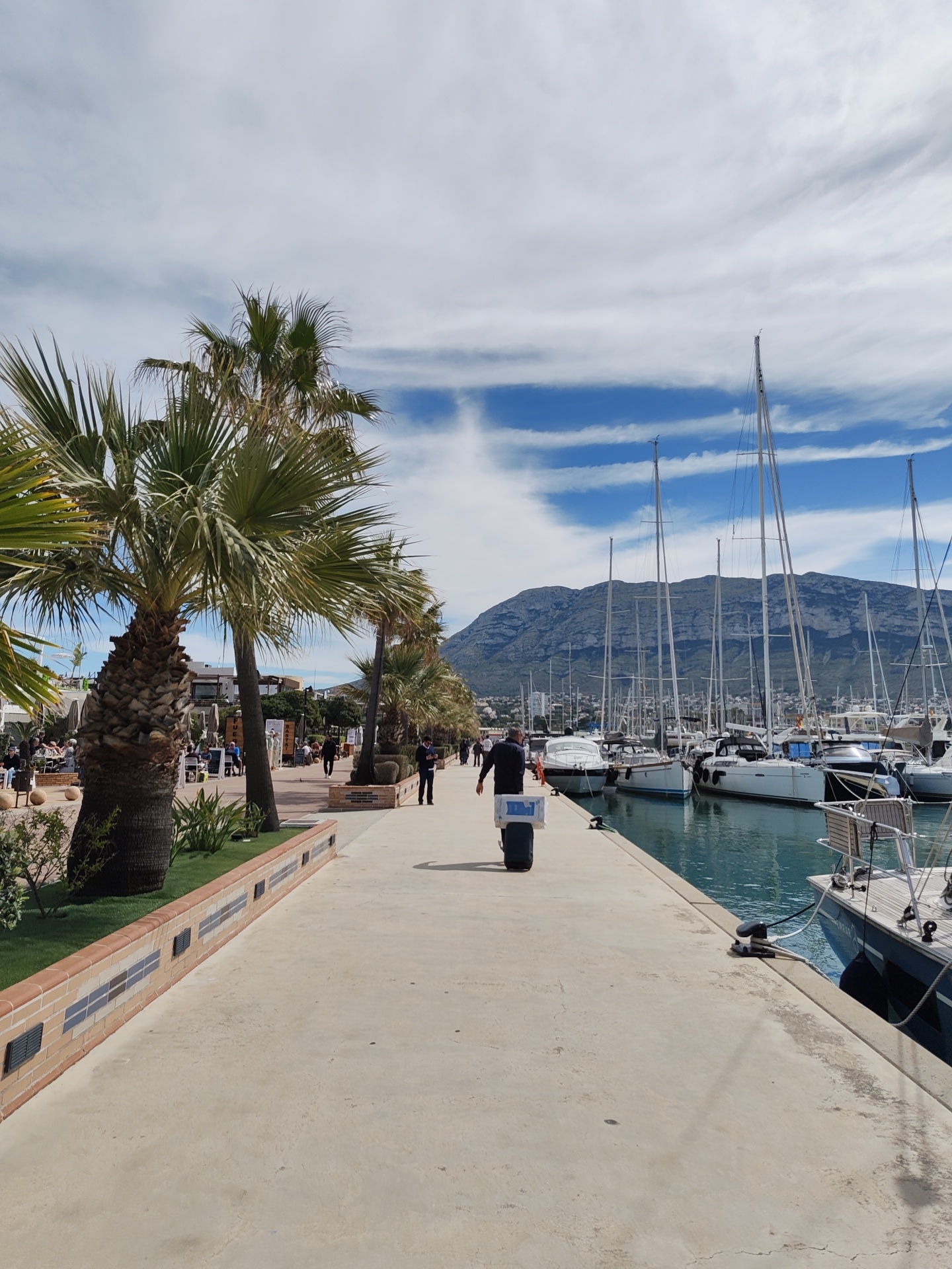 The image shows a view along a marina promenade in Dénia, Spain. Palm trees line the left side of the paved walkway. To the right, yachts and sailboats are docked along the water's edge. A tall mountain range forms the backdrop of the harbor. A man is seen walking away from the camera down the promenade pulling a suitcase. Other people are visible in the background walking along the promenade and sitting at a waterfront restaurant. The sky is partly cloudy with blue patches visible.