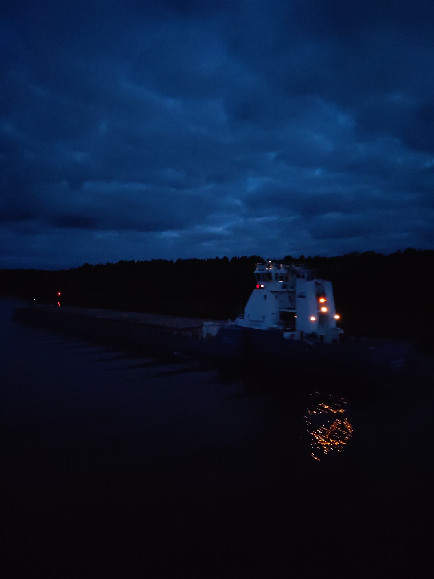 At night, a barge with a brightly lit tugboat attached moves on a river near Uzhla, Russia. A dark forest lines the riverbank in the background. The dark, cloudy sky is above. The lights from the tugboat reflect in the river.