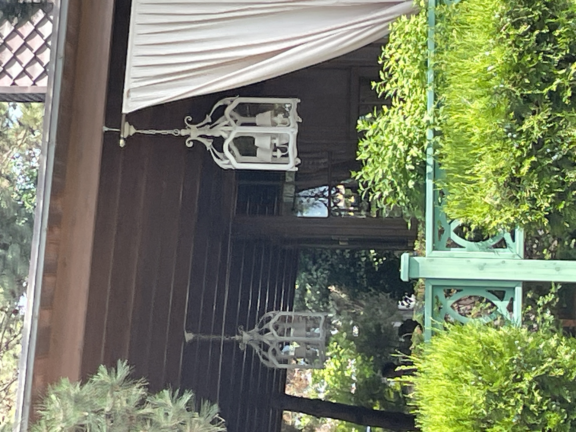 This image shows an outdoor scene, likely a patio or porch area. In the foreground, there is a bright green decorative fence with intricate cutouts. Behind the fence, there are lush green bushes and plants. Above the fence, suspended from the wooden ceiling of a structure, are two white lantern-style light fixtures. The fixture on the right is larger and more ornate, with a decorative top and multiple candle-style lights inside. The fixture on the left is smaller and simpler. To the right of the larger lantern, a light-colored curtain hangs. The background reveals wooden walls, windows, and foliage from trees. The scene appears to be well-maintained and suggests a pleasant outdoor living space. This image was captured in Anapa, Russia.