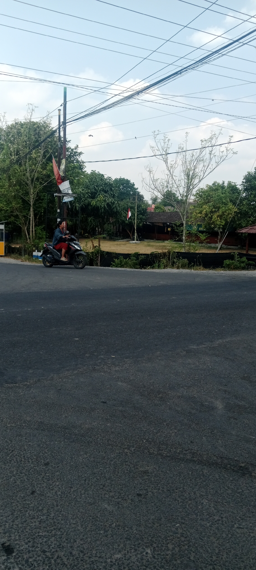 The image depicts a street scene in Maguwoharjo, Indonesia. In the foreground, a dark asphalt road occupies the lower half of the frame. In the middle ground, a person is riding a dark-colored motorcycle on the left side of the road. The rider is wearing a dark jacket and red pants. To the right of the road, there is a grassy area with trees and a building in the background. Two Indonesian flags are visible; one on a pole near the left side of the frame, and another smaller one further back on the right. Utility poles with numerous wires crisscross the sky, which is partly cloudy. The overall scene suggests a rural or suburban environment with typical infrastructure.