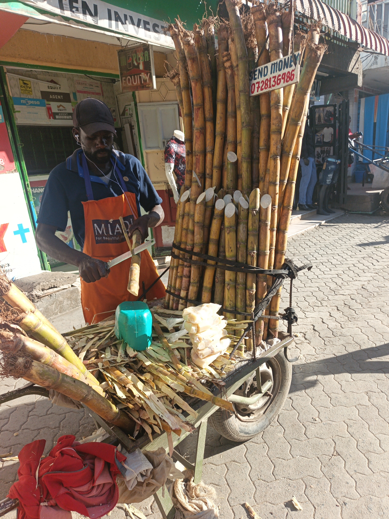 The image displays an outdoor street scene in Nairobi, Kenya, under bright daylight. A dark-skinned male vendor, wearing a navy blue shirt, an orange apron with "MIAD For Beautiful Life" text, a dark baseball cap, and white wired earphones, is actively peeling a segment of sugarcane with a large knife held in his right hand, while grasping another segment in his left hand.
He operates from a mobile, two-wheeled cart positioned on a patterned grey interlocking concrete block pavement. The cart carries a large, upright bundle of numerous yellow-brown sugarcane stalks, secured by black straps, forming a tall barrier on the right side of the frame. A white rectangular sign with red text reading "SEND MONEY 0728136986" is affixed to this sugarcane bundle. The cart's flatbed holds a pile of pale, peeled sugarcane segments, discarded sugarcane peels and leaves, several unpeeled sugarcane stalks, and a bright turquoise plastic container. A crumpled red garment and a light-colored cloth are visible on the front-left edge of the cart.
In the background, behind the vendor, is a building with a light-colored facade and green trim, featuring storefronts. Visible signage includes "EQUITY AGENT," "M-PESA," and "CCTV," confirming the Kenyan urban setting. A brown corrugated awning extends over the storefronts. Further back on the right, another storefront with a blue exterior is visible. Indistinct figures of other individuals and a mannequin in a shop window are present in the background. Distinct shadows are cast on the pavement due to the overhead sun.