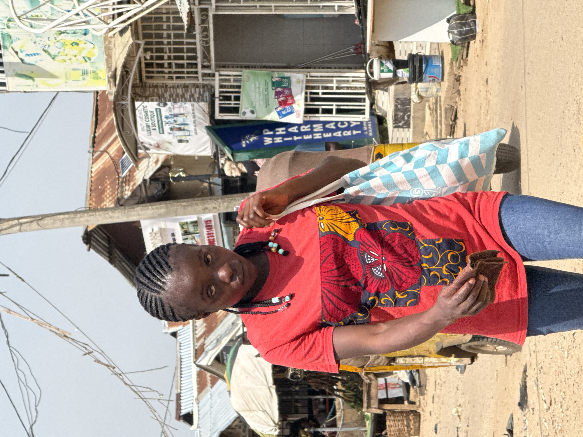 A young Black woman with braided hair is walking on a dirt road in Abuja, Nigeria. She is wearing a red t-shirt with a colorful floral pattern on the front and blue jeans. She is carrying a blue and white checkered bag in her right hand and a brown wallet in her left hand. Behind her, there are several market stalls with corrugated metal roofs, a yellow trishaw, and a sign for "White Heart Pharmacy." The sky is clear and bright, indicating a sunny day. The overall scene depicts a typical bustling street market in a Nigerian city.