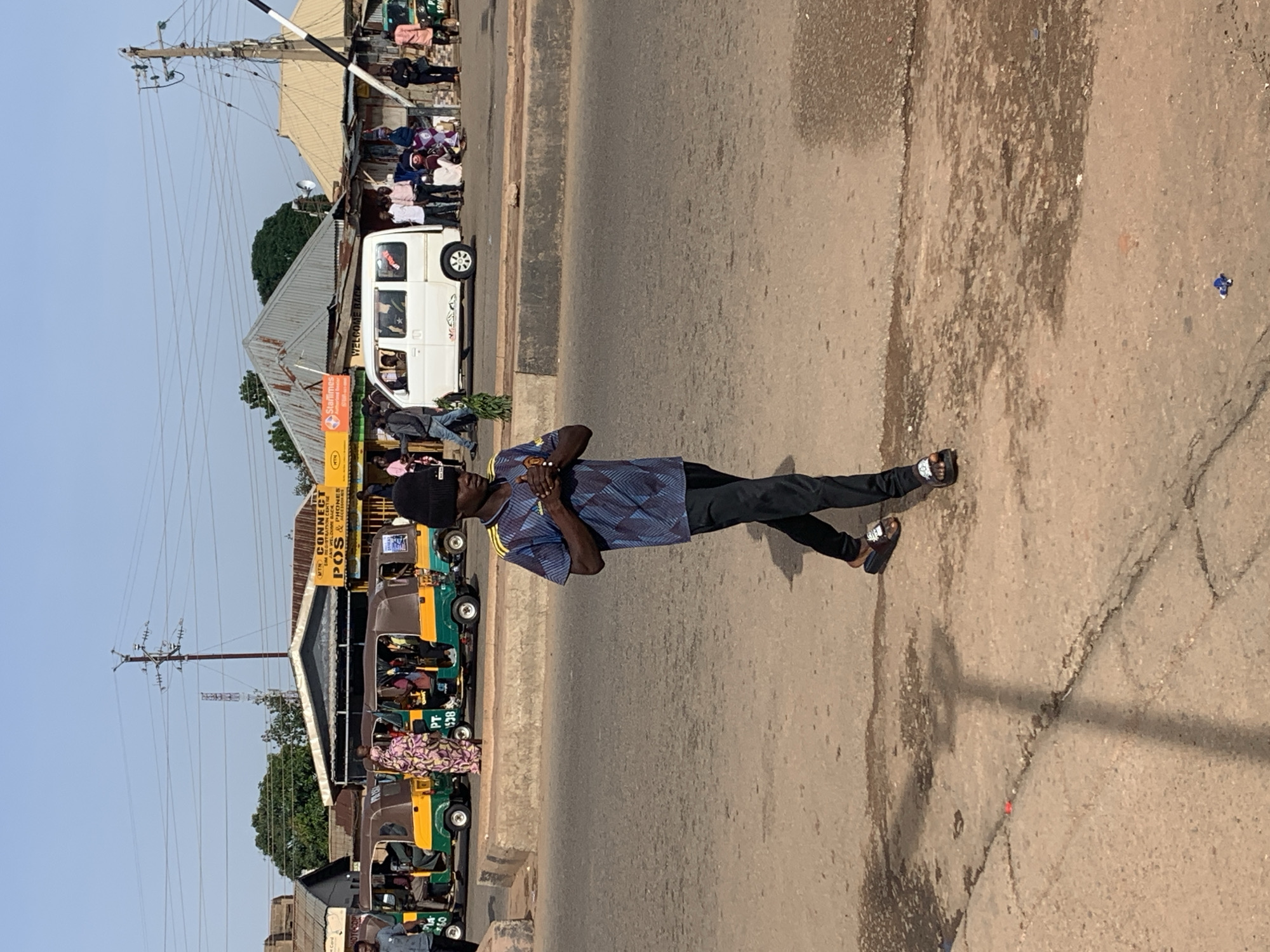 The image shows a street scene in Gura Top, Nigeria. A young man in a blue and black patterned shirt and black pants is walking across the street, holding his hands clasped in front of him. He is wearing black sandals. Behind him, several auto-rickshaws are parked or moving, with passengers visible inside. A white van is also present, as are other people standing on the sidewalk and near the vehicles. Several buildings with corrugated metal roofs line the street. Utility poles and power lines are visible in the background against a clear blue sky. The road surface appears to be asphalt, with some cracks and water stains.