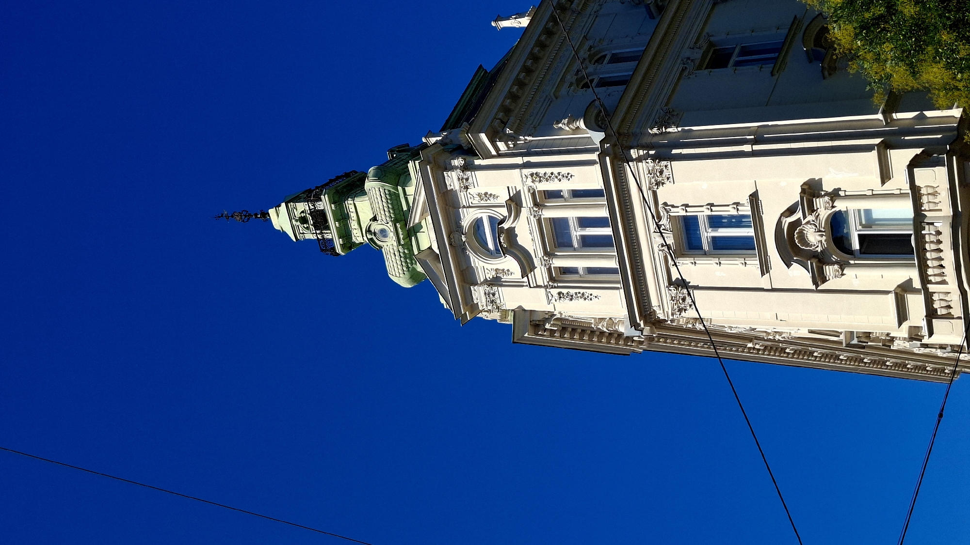 The image captures the upper facade of a cream-colored building with ornate architectural details, including intricate carvings and decorative window surrounds. The building features a green, domed turret topped with a dark metal finial and a small balcony with wrought-iron railings. The bright blue sky serves as a backdrop, with two dark utility wires diagonally crossing the frame. A small portion of lush green foliage is visible in the lower right corner. The building's style suggests it is located in a historic European city. This building is located at Kohlmarkt 12, Vienna, Austria.