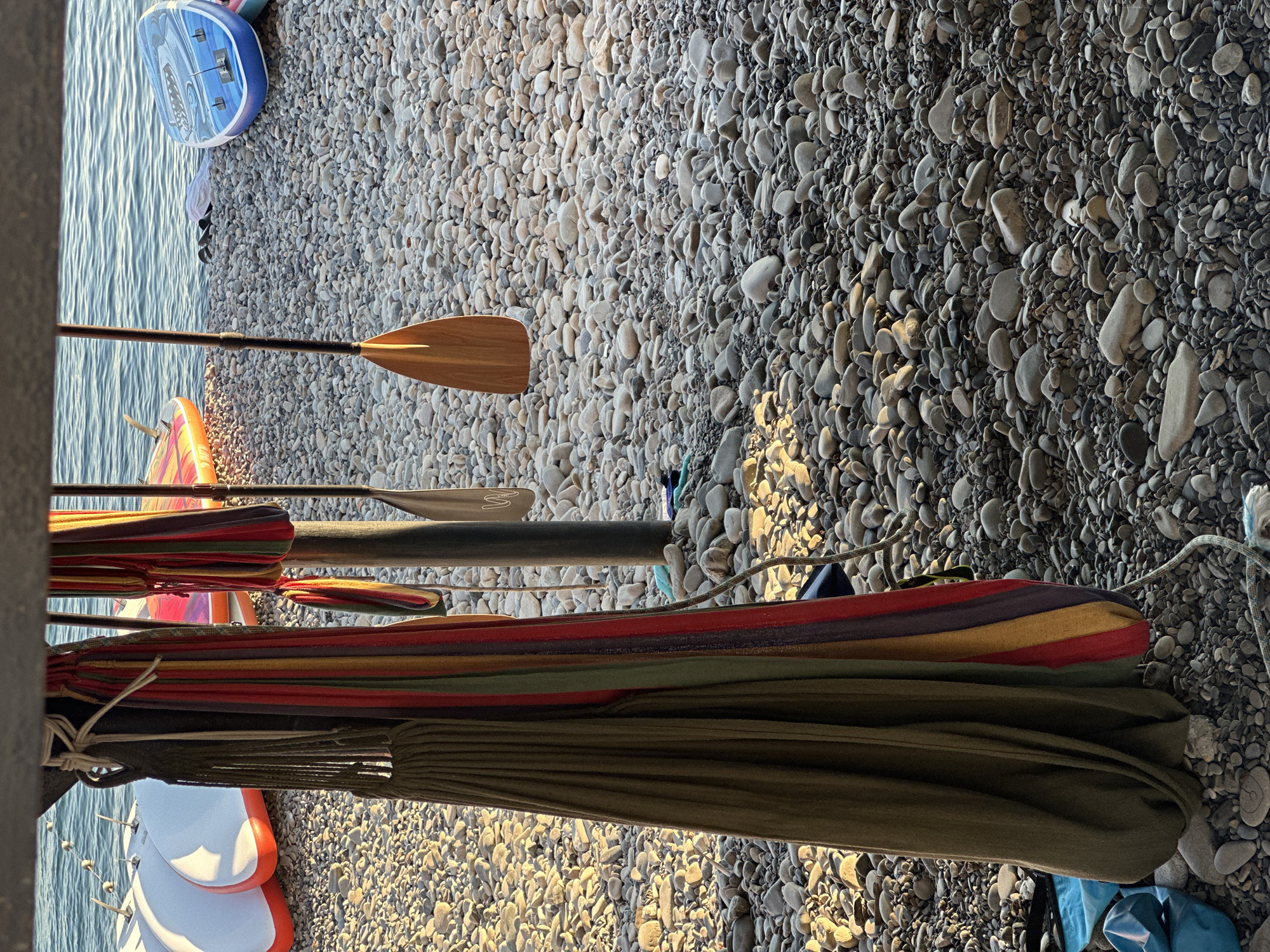 The image depicts a rocky beach scene with several stand-up paddleboards and a hammock displayed. The water in the background suggests a coastal location. The paddleboards are of various colors, including white, orange, and blue. A wooden paddle hangs from a vertical pole, and a multi-colored hammock is also suspended nearby. The ground is covered in small, smooth stones. The presence of paddleboards and a hammock implies a recreational area, possibly a beach resort or a park. The overall lighting indicates it is daytime, with sunlight casting shadows on the ground. The scene appears to be set in a natural outdoor environment.