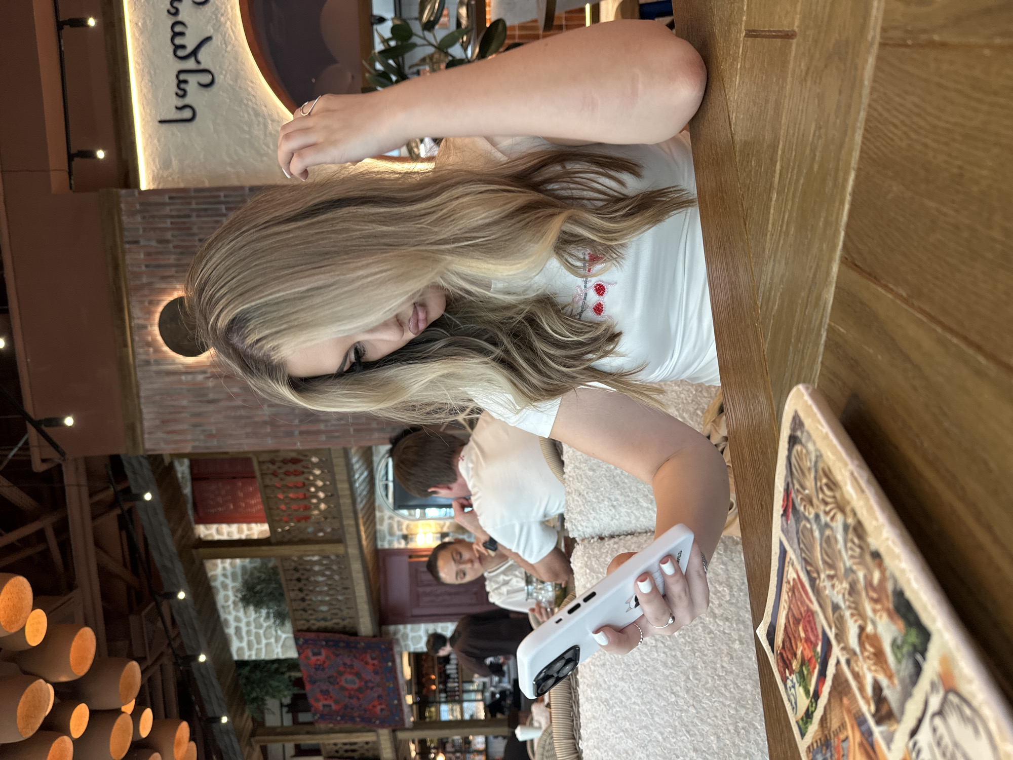 The image features an indoor scene, likely a restaurant or cafe, with a young woman as the primary subject. She has long, blonde hair and is viewed from her left side, looking down at a white smartphone held in her right hand. Her left arm rests on a light brown wooden table. She is wearing a white top with red embroidered details, possibly depicting strawberries. Her fingernails are painted white, and she wears rings on her fingers. The white smartphone has a prominent camera module and is encased in a white cover.
The background reveals a warm and culturally themed interior, characterized by exposed brick walls, textured walls, and various decorative elements. A significant detail is a stylized inscription in Georgian script on a textured wall, reading 'ქართული' (kartuli), which means 'Georgian.' Other patrons are visible in the background, including a man in a white t-shirt. The decor includes cylindrical hanging light fixtures, string lights, and patterned rugs or tapestries adorning the walls. On the table next to the woman, there is a decorative plate or menu with mosaic-like images. The overall ambiance suggests a casual gathering or meal. The time of day is not explicitly clear, but the interior is well-lit by artificial lights.