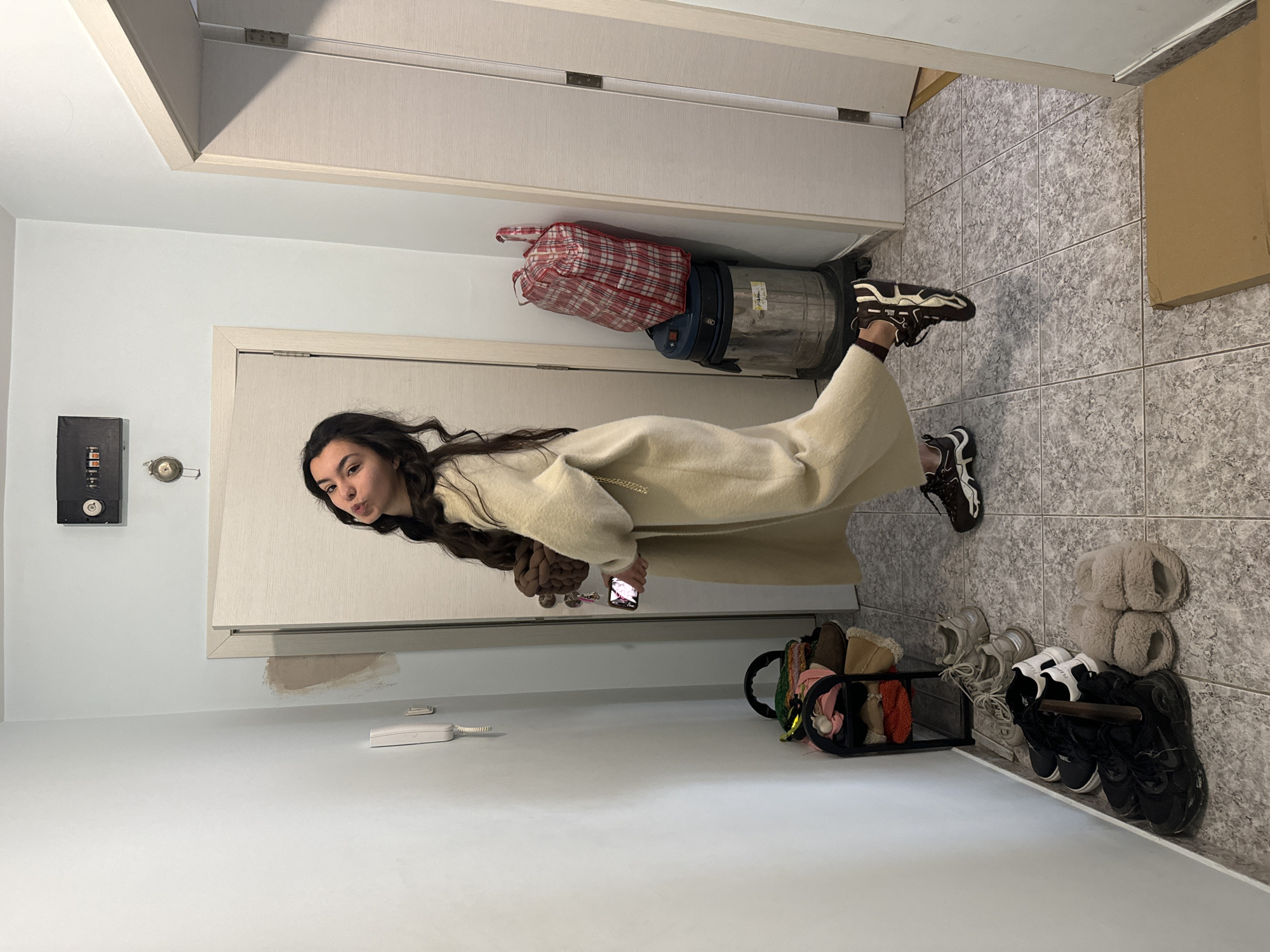 A woman in a long, beige coat and sneakers stands in a hallway. She is holding a phone in her right hand. To the left, there is a wall with a fuse box and a telephone. In front of her is a shoe rack filled with various shoes and slippers. Behind her, a vacuum cleaner with a red and white checkered bag on top rests against the wall. The hallway has gray tile flooring and a beige door. The location is likely in a residential building in Varna, Bulgaria, given the architectural style and interior design.