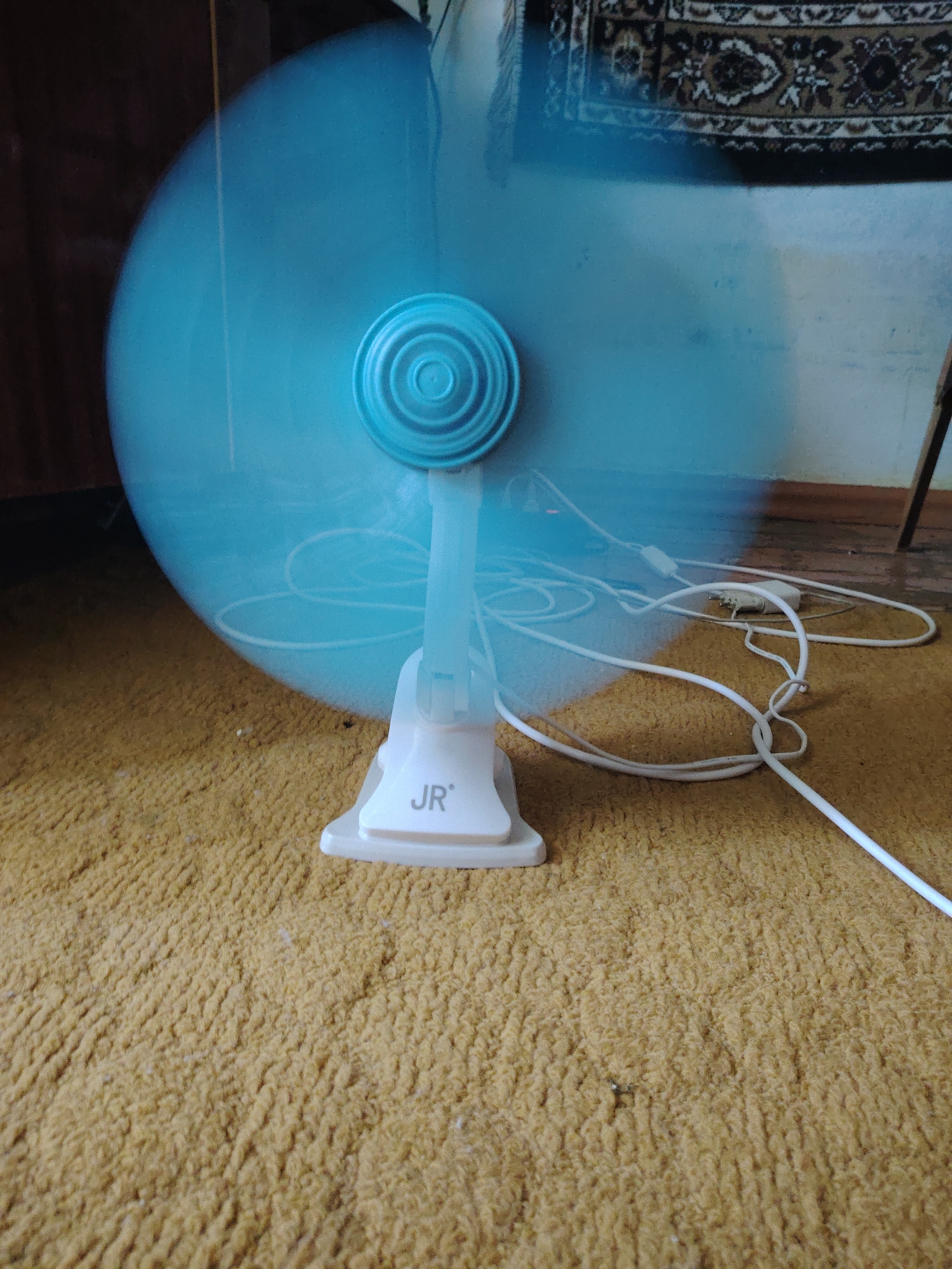 An indoor scene, likely a residential interior in Chirchik, Uzbekistan, features a close-up view of a blue electric fan with blurred blades indicating it is in operation. The fan is positioned on a textured, mustard-yellow carpet. The base of the fan has a white clip and bears the "JR" logo. A white power cord is visible, extending from the fan and lying across the carpet, connecting to an adapter plug. In the background, a dark wooden furniture piece is on the left, and a patterned rug or tapestry hangs on the wall to the right. The overall lighting is dim, with light emanating from the fan's blades.