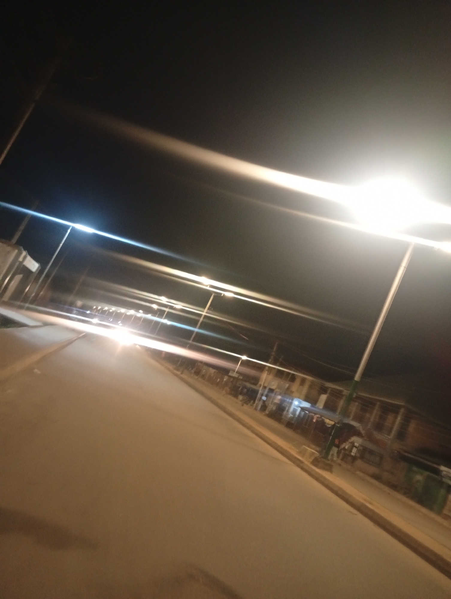 An outdoor, full shot of a street in Bukuru, Nigeria at night. The street is paved and appears to be an empty road with streetlights lining the side. The streetlights are tall and thin with white lights. The buildings on the side of the street are dark, but lights can be seen inside some of them. The sky is black with light streaks from the streetlights. The camera angle is angled downward, showing the road ahead.