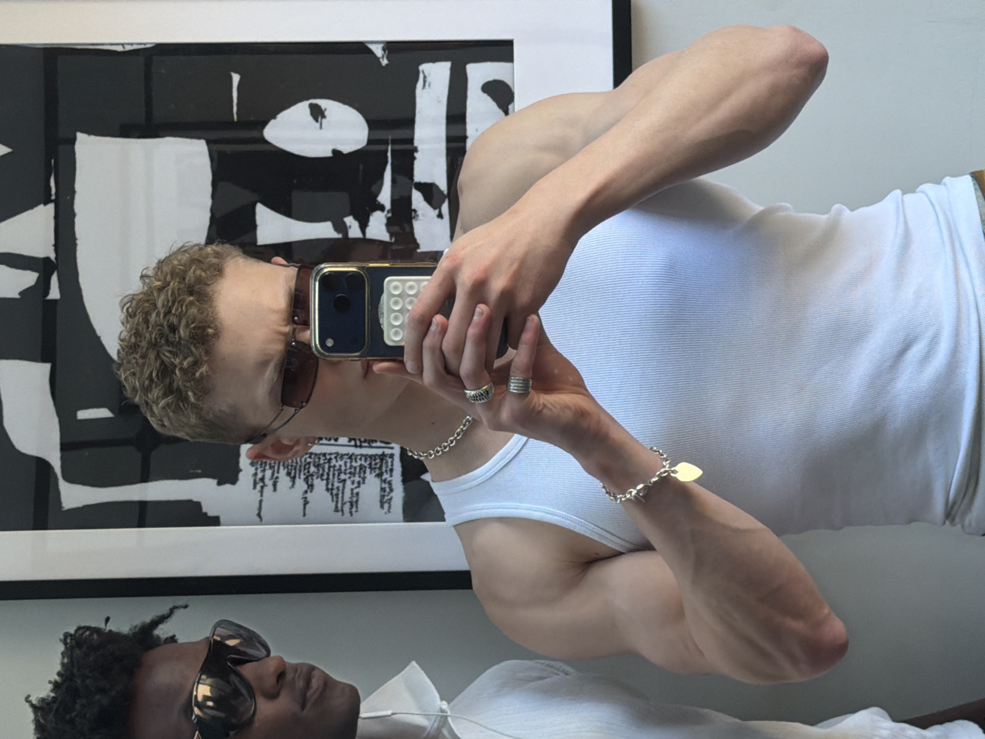 A fair-skinned man with curly blond hair is taking a photograph of himself in a mirror. He is wearing a white tank top, sunglasses, a silver chain necklace and bracelet, and rings. He is holding a cell phone with a case. The reflection shows another man, who has dark skin, wearing sunglasses and a white shirt. Behind the men is a framed black and white abstract print. The location is unknown but hypothesized as London, United Kingdom.