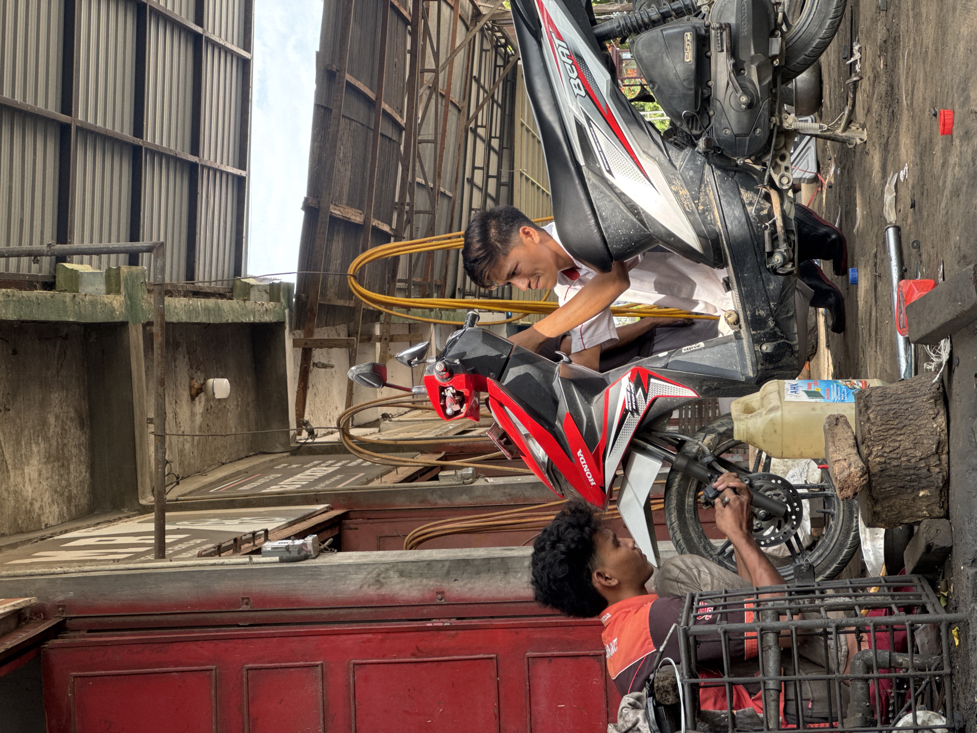 In Meunasah Pupu, Indonesia, two young men are working on motorcycles in an outdoor workshop. The workshop appears to be located in front of a building with a red door and a corrugated metal roof supported by metal beams. Sunlight illuminates the scene.
One man, wearing an orange and black shirt, is crouched down and appears to be focused on the front wheel of a red and black Honda motorcycle. He is holding tools and working on the braking mechanism. A metal cage-like structure is beside him, holding some items.
The other man, wearing a white shirt, is standing and also working on a motorcycle. This motorcycle is primarily black with red and silver accents. He is holding a tool near the handlebars. He is wearing black shoes.
The ground is dirt and debris, with scattered tools and motorcycle parts. A yellow jerrycan with a blue label is visible near the front wheel of the red and black motorcycle. Wooden blocks are also present on the ground. In the background, yellow hoses are coiled and hanging. The overall impression is of a busy, functional repair shop.