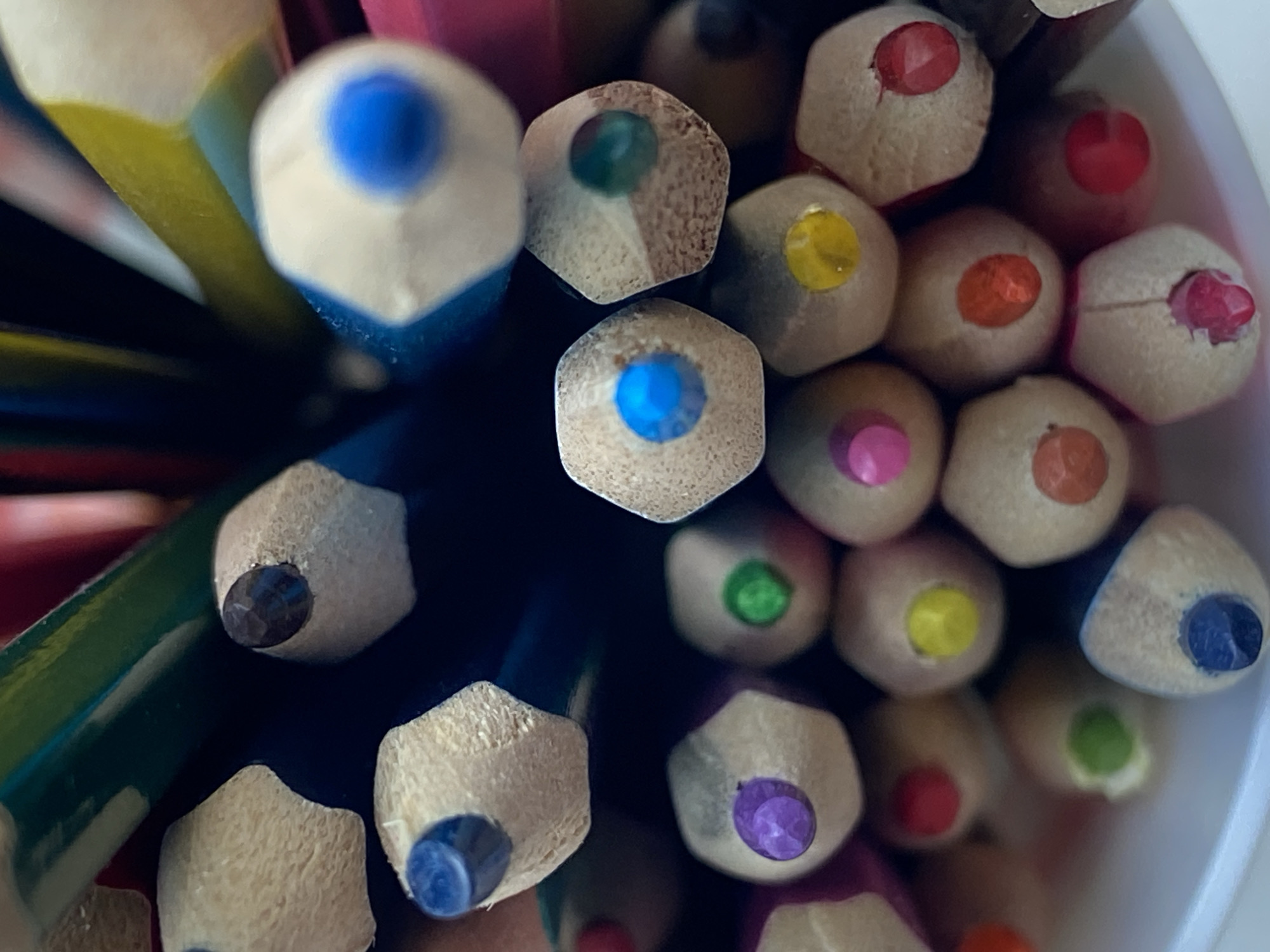 A close-up, overhead shot shows a cluster of sharpened colored pencils in varying colors. The pencils are packed tightly together, with their pointed tips facing downwards and out of focus towards the bottom of the frame. The tips of the pencils are visible, each displaying a different color, such as blue, green, purple, pink, yellow, and red. The wood grain of the pencils is textured and detailed. The lighting is bright and highlights the colors and textures of the pencils. The background is dark and blurry, with hints of other colored pencils continuing the composition. The image appears to be taken indoors, possibly in a craft or art supply setting. There are no people, animals, or any specific city or environmental details visible in the image.
