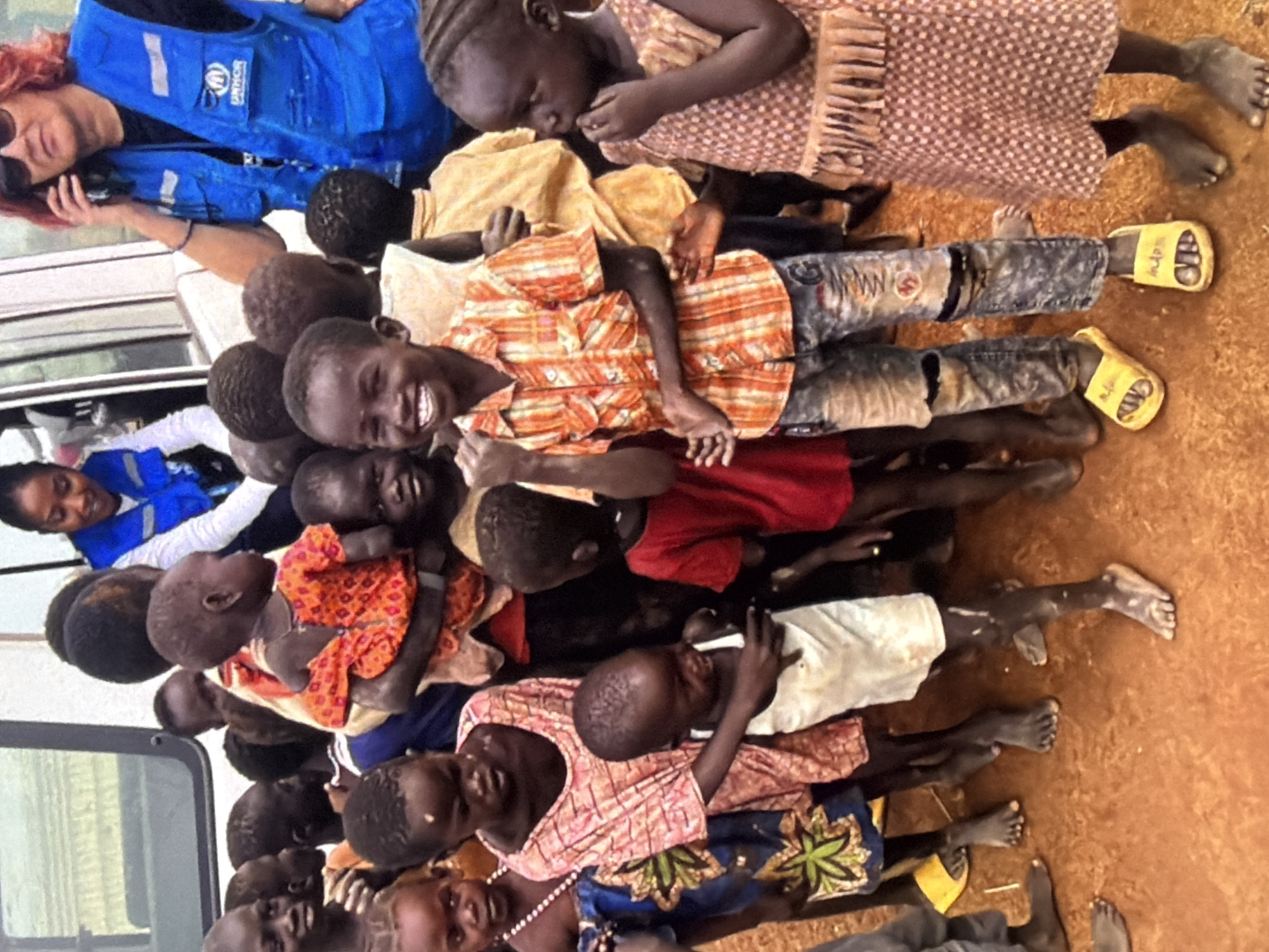 The media file shows a group of people, predominantly children, lying down together on a reddish-brown, dusty surface. The scene appears to be outdoors, with the ground suggesting a dry, possibly rural or encampment setting. The lighting suggests it is daytime.
The individuals are arranged in a line, with some appearing to be sleeping or resting. The children are of various ages and are dressed in colorful clothing, including patterned shirts and dresses. Some are wearing yellow flip-flops. One child is being held by another.
In the background, a person wearing a blue vest with the UNHCR (United Nations High Commissioner for Refugees) logo is visible, along with parts of a white vehicle. This suggests a context related to humanitarian aid or refugee support.
The expressions on the faces of the visible children range from peaceful to mildly alert. The overall mood is one of communal gathering and rest in a possibly challenging environment. There is no clear indication of a specific event, but the presence of UNHCR personnel and the gathering of people imply a situation where aid is being provided or managed. No other text is discernible apart from the UNHCR logo.
