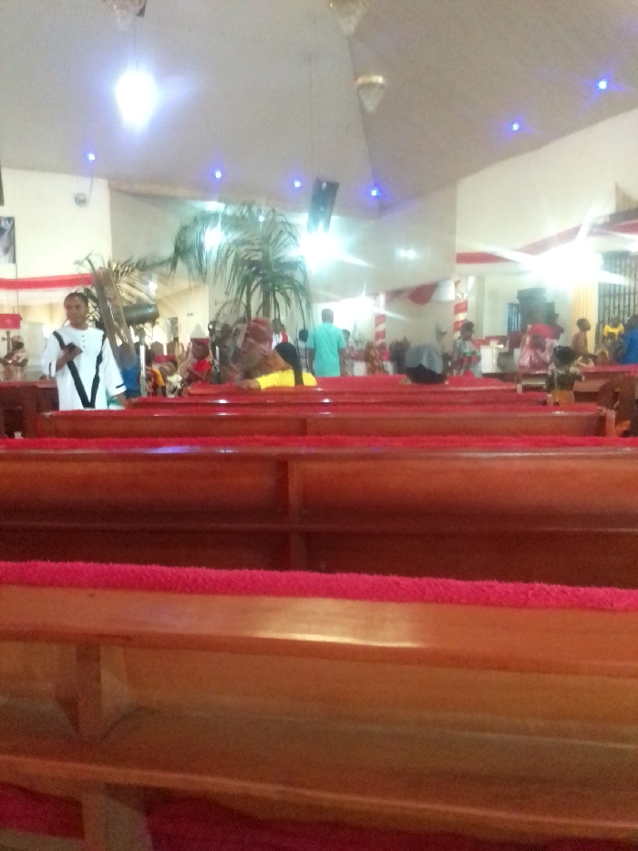 The image shows a view from the back of a pew inside a church. There are several rows of pews in front of the viewer, made of wood and padded with red fabric. In the background, people can be seen sitting in the pews and standing in the aisle. There is a large palm tree in the middle of the room, and the ceiling has several bright white lights. The image was taken inside a church in Auchi, Nigeria, located at 7.068 latitude and 6.269 longitude.