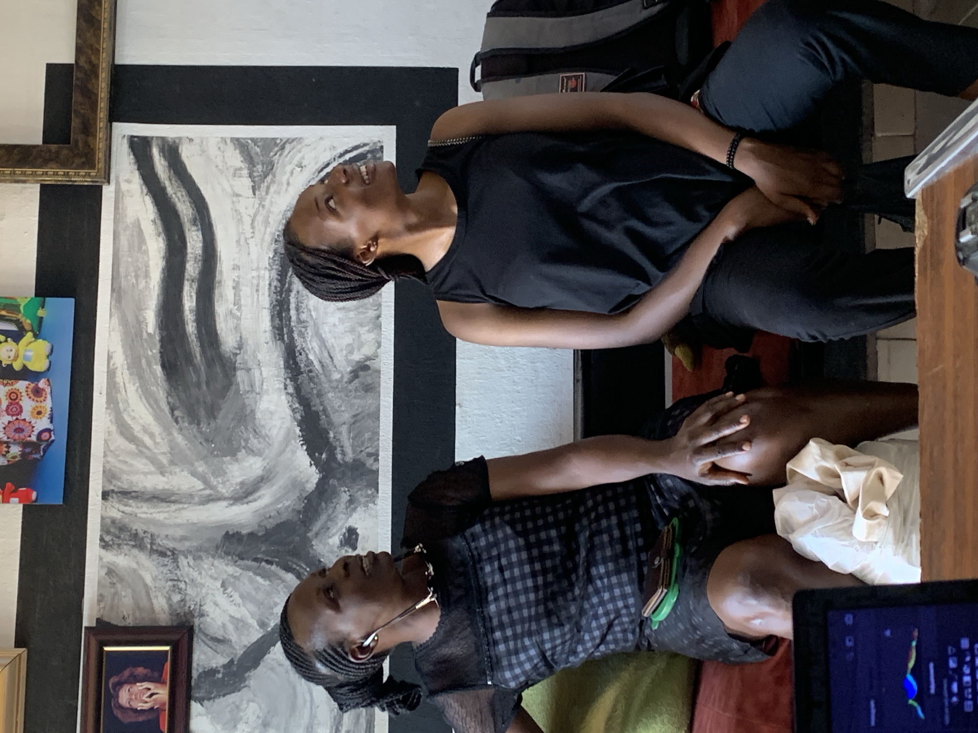 Two Black women are indoors. The woman on the top is wearing a black tank top and black pants. She has her hair braided and is smiling. The woman below her is wearing a black and gray patterned dress with a sheer top and a necklace. Her hair is also braided. Behind them is an abstract painting of gray and white swirls on a wall with black and white stripes. To the left of the woman on the bottom is a brown framed portrait. To the right is a laptop. Jos, Nigeria is not evident in the image.