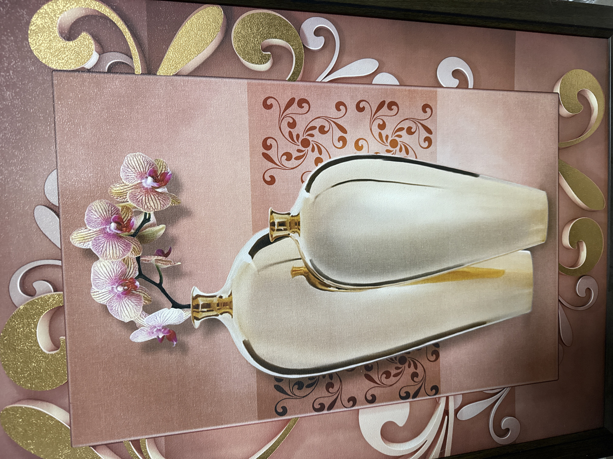 The image shows a framed artwork with a complex design. The artwork features two stylized, elongated vase shapes laid on top of each other, colored in shades of beige and gold. Beside the vases is a cluster of pink and white orchids. The background of the artwork is composed of alternating brown and beige squares, adorned with floral patterns in brown and gold. Decorative, ornate swirls in gold and white border the central rectangular design. The artwork is contained within a dark brown frame.
There is no information provided in the image that indicates the artwork's location in Votkinsk, Russia, or any other specific location.
