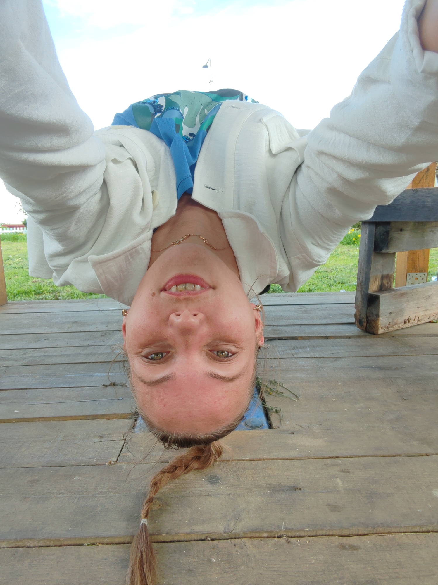 The image shows a person hanging upside down, with their head positioned over a gap in a wooden platform. The person is wearing a white shirt or jacket and a blue top underneath. They have a braid in their hair. The location is Чухлэм, Russia. The background includes a grassy area and a glimpse of a structure. A wooden bench or table is visible to the right.