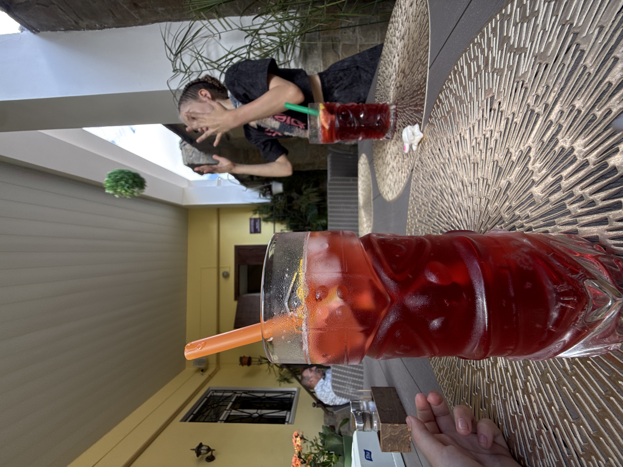 The image depicts an outdoor seating area, possibly a cafe or restaurant patio. In the foreground, a tall, tiki-shaped glass filled with a red beverage and ice is prominently displayed. An orange straw extends from the drink. To the left, a person's hand with painted nails rests on the table. In the mid-ground, another tiki glass with a red drink and a green straw is visible. Further back, a young person with braided hair is seen interacting with a small, green spherical object, which is in mid-air, suggesting they are playing or tossing it. The background reveals a building with yellow walls, an open doorway, and what appears to be outdoor furniture and foliage. The lighting suggests it is daytime.