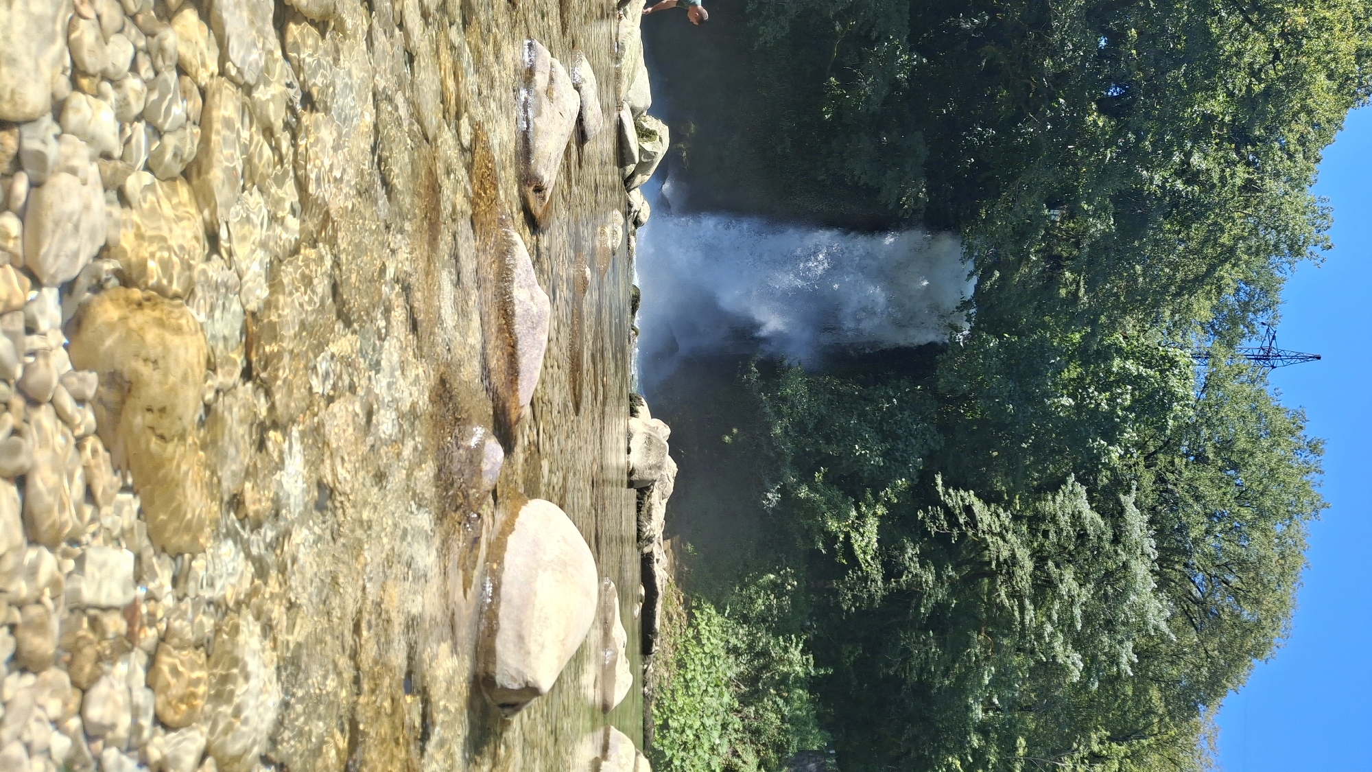 In Gachedili, Georgia, a low-angle shot captures a natural scene featuring a cascading waterfall. The waterfall, framed by lush green trees and foliage, plunges into a pool of water. In the foreground, the crystal-clear water reveals a rocky riverbed adorned with smooth, light-colored stones of various sizes. Sunlight glints off the water's surface, creating a shimmering effect. A person, partially visible on the left side of the frame, is standing near some rocks by the water's edge, facing the waterfall. In the background, the dense green canopy of trees dominates the landscape, with a clear blue sky overhead. A tall, dark industrial structure, possibly a pylon or tower, is visible above the treeline on the right side of the image.