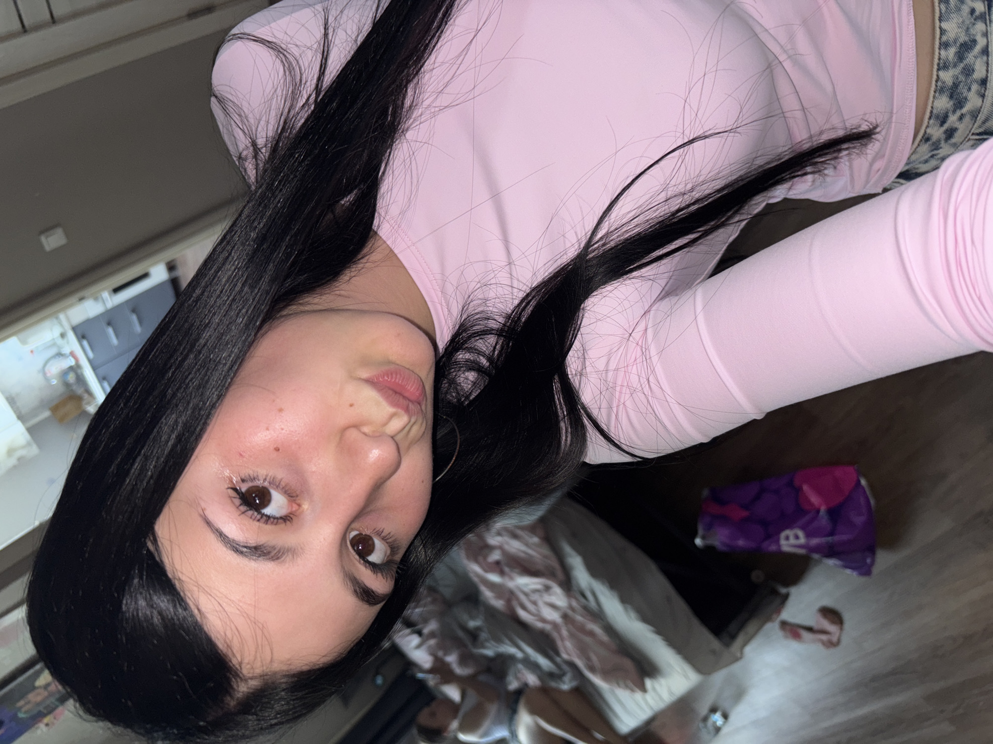 A young woman with long, dark hair and brown eyes is captured in a selfie taken indoors. She is wearing a pale pink long-sleeved shirt and appears to be pouting her lips. In the background, another person is visible lying on a bed with pink and grey bedding. Various objects, including a purple bag with a heart design and what appears to be a piece of clothing, are scattered on the wooden floor. The environment suggests a domestic setting, possibly a bedroom or living area, though no specific details about the city of Chaikovsky, Russia, are discernible from the image.
