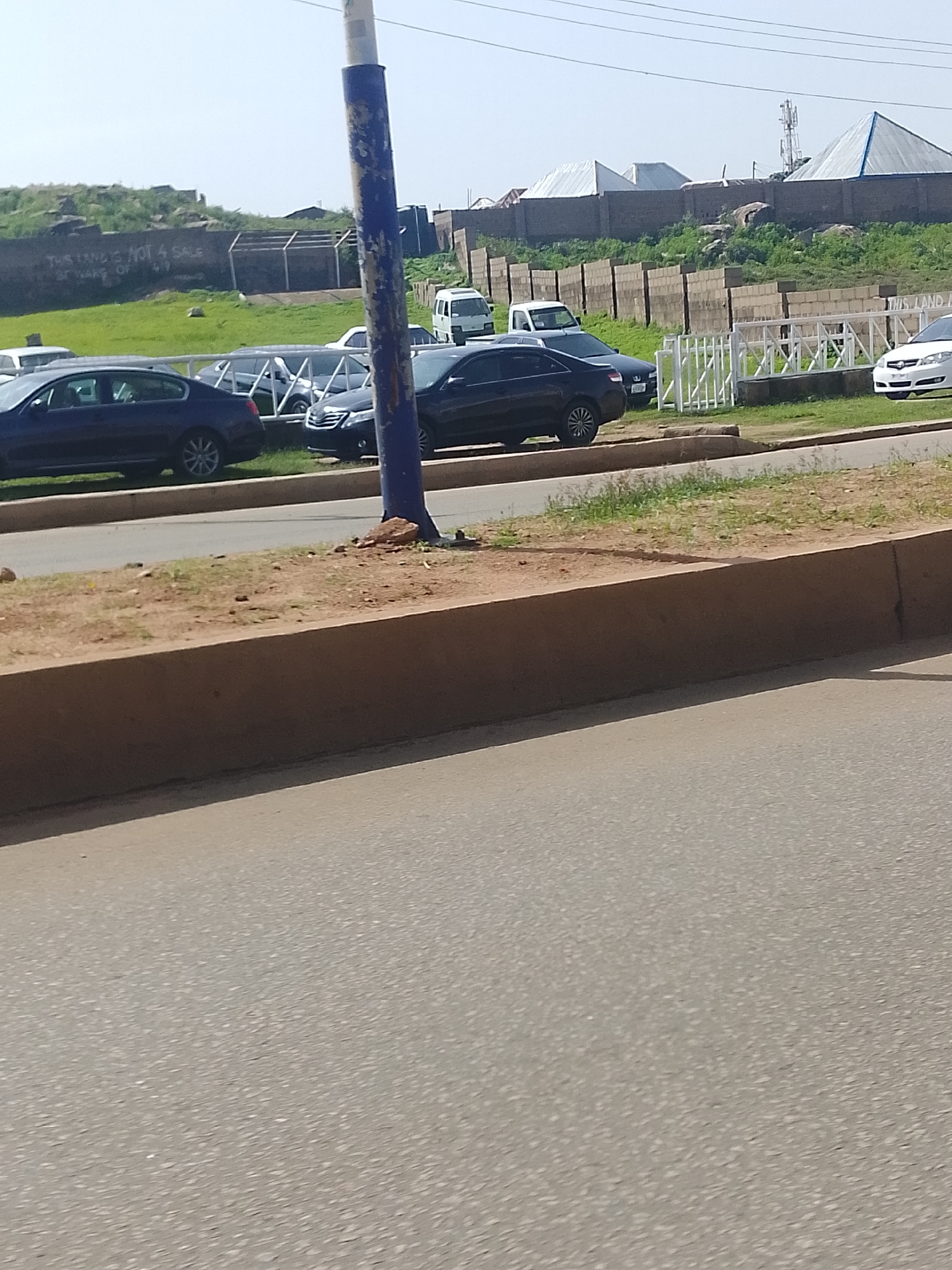 In Bukuru, Nigeria, a dark-colored sedan is parked on a paved road adjacent to a raised earthen embankment. Behind the embankment, a wall constructed of concrete blocks encloses an area with green grass and scattered rocks. Further in the background, several buildings with distinctive peaked roofs are visible, along with a white fence and a tall antenna structure. Another dark sedan is parked in front of the white fence. To the left of the sedan, a section of a wall with graffiti is present. The sky above is clear and light blue. The perspective suggests the image was taken from a moving vehicle.