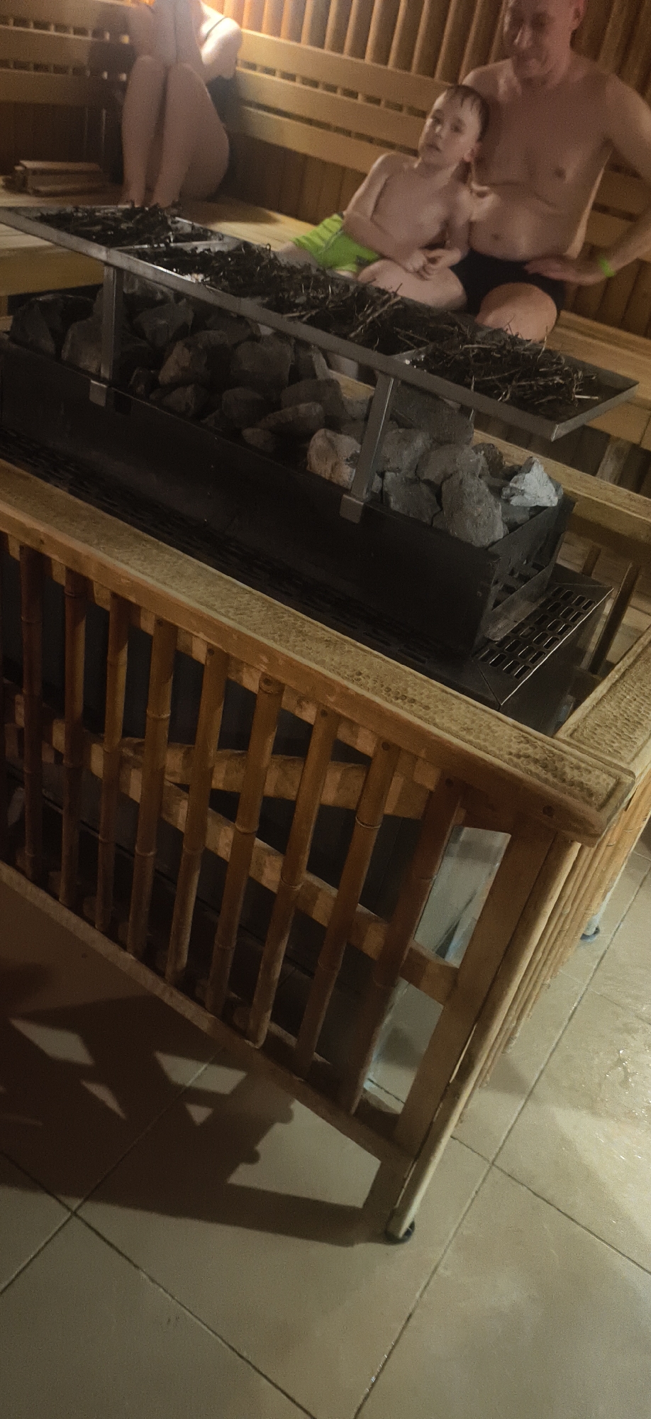 This image depicts an indoor scene within what appears to be a traditional Russian banya or sauna. The setting features wooden benches, a prominent sauna heater, and tiled flooring.
In the foreground, a substantial sauna stove or heater is visible. It's a dark metal unit filled with numerous gray-black sauna rocks. Above the main rock bed, a metal grate or shelf holds a large quantity of dried branches or twigs, commonly known as "veniks" in Russian saunas, typically used for whisking during steam sessions. A light-colored wooden safety barrier, constructed with vertical and horizontal slats, partially encloses the heater, providing a protective element. The floor beneath is covered with light, rectangular tiles, some appearing damp.
Several individuals are present, suggesting a family or group enjoying the sauna. On the right side, an adult man, wearing dark swimming trunks, is seated on a wooden bench. Beside him, a young child, wearing light green swimming trunks, is also seated, looking directly towards the camera with a neutral expression. Further to the left and slightly obscured by the foreground, another person, likely an adult, is seated on a higher wooden bench. Only their legs and lower torso are clearly visible, indicating they are also in swimwear.
The lighting in the room is warm and somewhat dim, contributing to a relaxing atmosphere. Shadows from the wooden barrier are cast on the tiled floor. The presence of veniks strongly hints at a traditional Russian banya experience, fitting with the location context of Votkinsk, Russia. No visible text is present in the image.