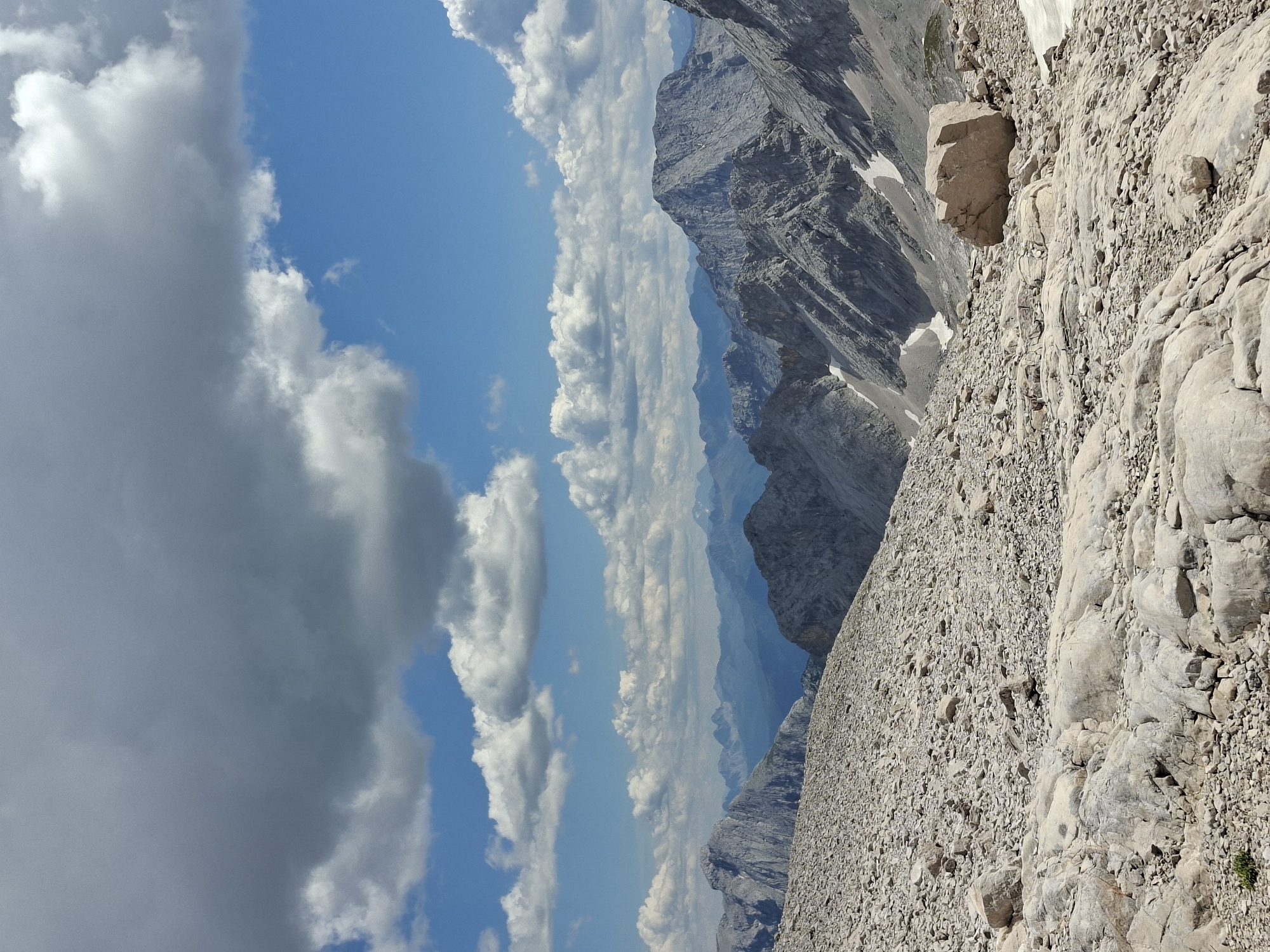 The image depicts a panoramic view of a rocky, mountainous terrain under a partly cloudy sky. The foreground is dominated by a dense scattering of light-colored rocks and boulders of various sizes, with larger, smoother rocks in the immediate foreground. In the mid-ground and background, a series of rugged mountain ranges stretch across the horizon, with jagged peaks and steep slopes. Some patches of snow are visible on the shaded sides of the mountains. The sky is a vibrant blue, punctuated by large, fluffy white clouds and darker, more ominous grey clouds in the upper portion of the frame. There are no visible people or structures in the image. The scene is indicative of a high-altitude, alpine environment, likely in the Bavarian Alps region of Germany, given the reference to Grainau.