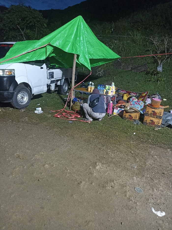 The nighttime image depicts a makeshift outdoor setup in Kecamatan Pujut, Indonesia, suggesting a temporary roadside stall or encampment. In the foreground, a person is seen crouching on a grassy patch, facing away from the viewer. They are dressed in a grey top, possibly with a hood, and light-colored pants, with a black fanny pack or small bag slung over their shoulder. They appear to be attending to a small, blue gas cylinder (LPG tank) positioned on the ground, surrounded by red straps or ropes.
To the left, a white pickup truck, resembling a Suzuki Carry or similar light commercial vehicle, is parked. A bright green tarp serves as a canopy, extending from the truck's roof to provide shelter over the setup, supported by a wooden pole. Underneath this tarp, to the right of the crouching person, a small folding table is laden with various items: multiple plastic bottles of water, several cups, and packaged goods that appear to be instant noodles or snacks, along with a thermos.
Beyond the table, on the grass, are personal belongings such as blankets and bags. Several cardboard boxes are stacked, with the "Kopiko" brand clearly visible on some of them. The ground transitions from a dirt path in the lower foreground to grass around the setup, with some scattered litter like plastic wrappers. In the background, a dark, vegetated hillside rises, indicating a non-urban, possibly rural or mountainous, setting. The scene is illuminated, suggesting artificial lighting is in use, given the dark sky. The overall impression is one of practicality and resourcefulness for a temporary stop or vendor activity.