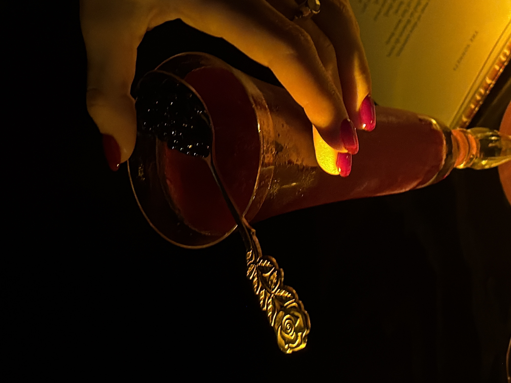 A close-up, dimly lit image captures a hand holding a cocktail glass in what appears to be an upscale evening setting in Đà Nẵng, Vietnam. The hand, with manicured nails painted in a vibrant fuchsia-pink, gently grips the glass, highlighted by a warm, golden light that casts dramatic shadows against the dark background.
Inside the tall, clear glass, a reddish-amber beverage is visible, garnished with a bright yellow lemon or citrus peel on the rim. A small, ornate spoon, featuring a distinctive rose design on its handle, rests within the drink. The spoon holds a serving of small, black, spherical garnish, resembling caviar.
The background is predominantly dark, emphasizing the illuminated hand, glass, and its contents. A light-colored menu or card is partially visible in the upper right, appearing blurred and upside down, with faint text that seems to include "THE SOMMELIER." The overall ambiance suggests a sophisticated and intimate experience at a bar or restaurant during nighttime.
