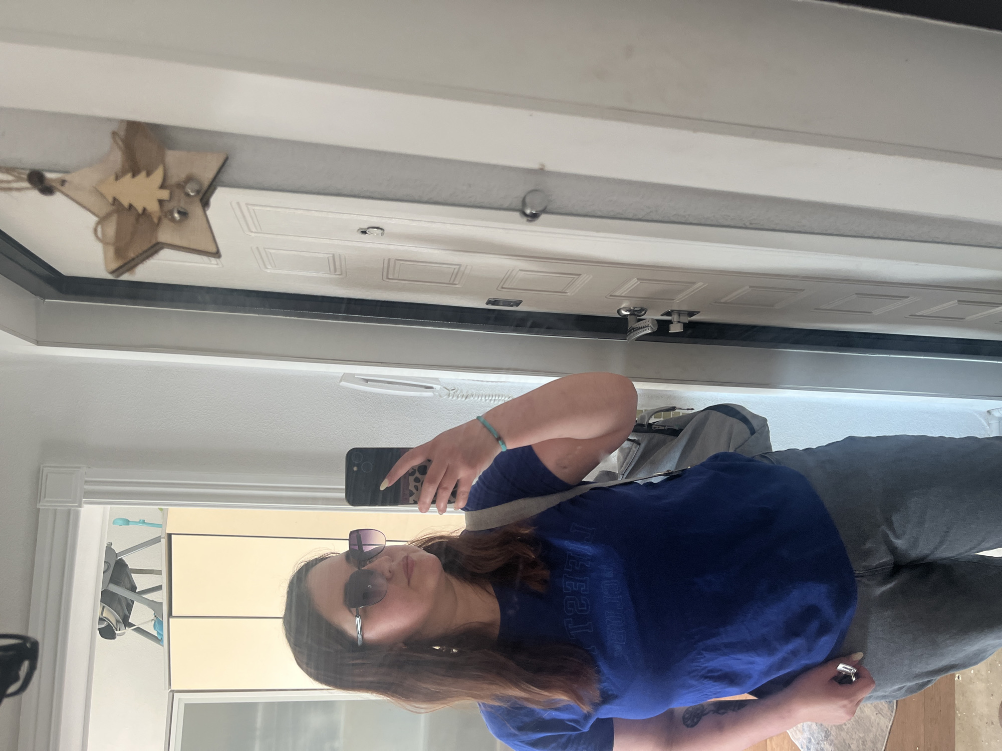 A young woman is taking a mirror selfie in a hallway. She is wearing a blue t-shirt, gray pants, and sunglasses. She is holding a smartphone in her right hand, and a gray backpack is slung over her left shoulder. In the background, a white door with a wooden Christmas ornament is visible. The hallway appears to be in an apartment building, with a closet and a crib visible in the distance. The woman is looking at her phone, and the photo is taken from a slightly low angle.