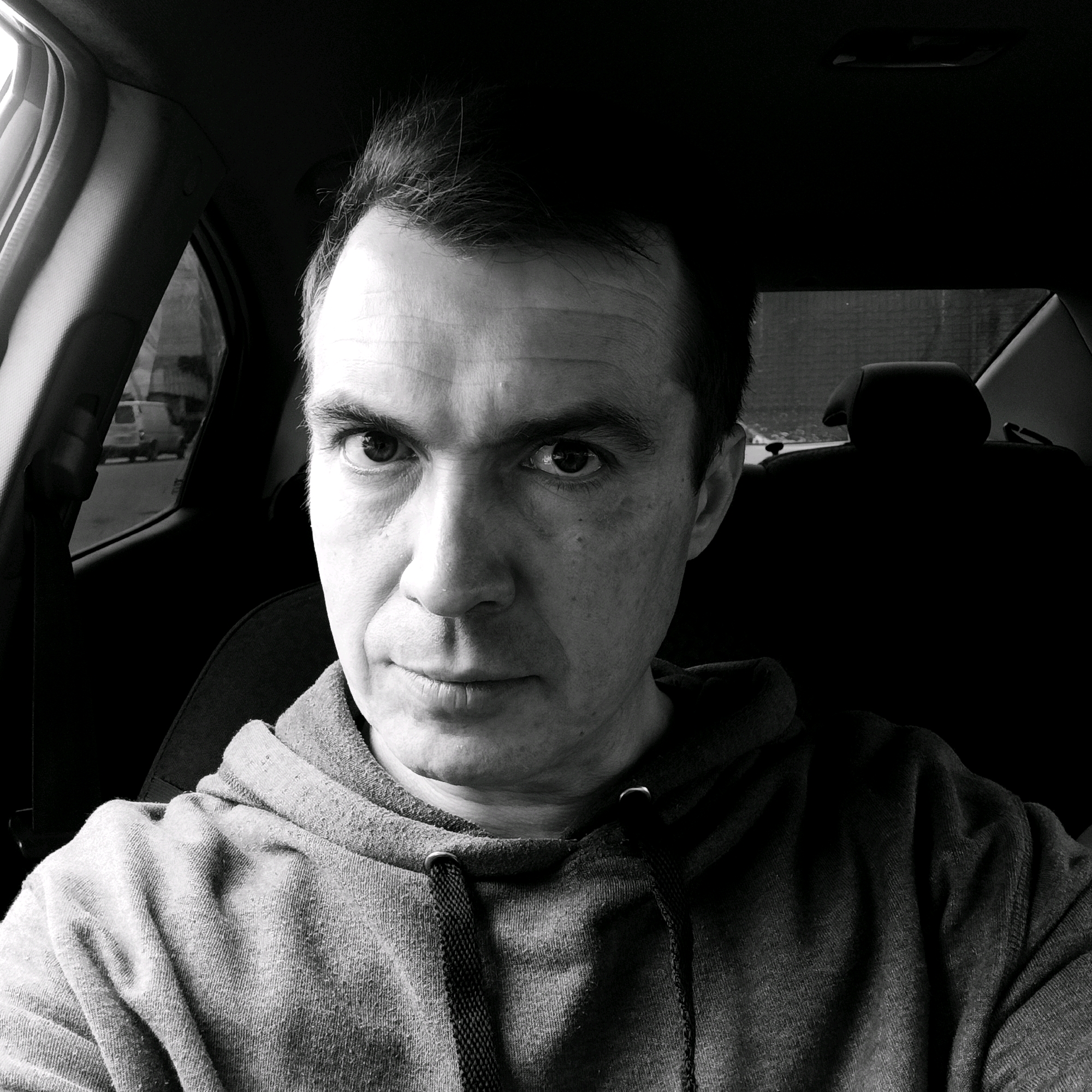 A black and white, close-up, medium shot, selfie style photograph shows a man sitting in the driver's seat of a car. The man is looking directly at the camera with a neutral expression. His hair is dark and appears styled upwards. He is wearing a dark hooded sweatshirt with drawstrings visible around the neckline. The interior of the car is visible, including the headrest of the seat behind him and a portion of the car's interior framing the windows. Light is coming from the side, highlighting the man's face and the texture of his sweatshirt. The background outside the car windows is blurred, but appears to be an urban environment with some buildings and vehicles. The photograph appears to have been taken during the daytime. No specific location in Nizhny Novgorod, Russia, can be identified from the image.