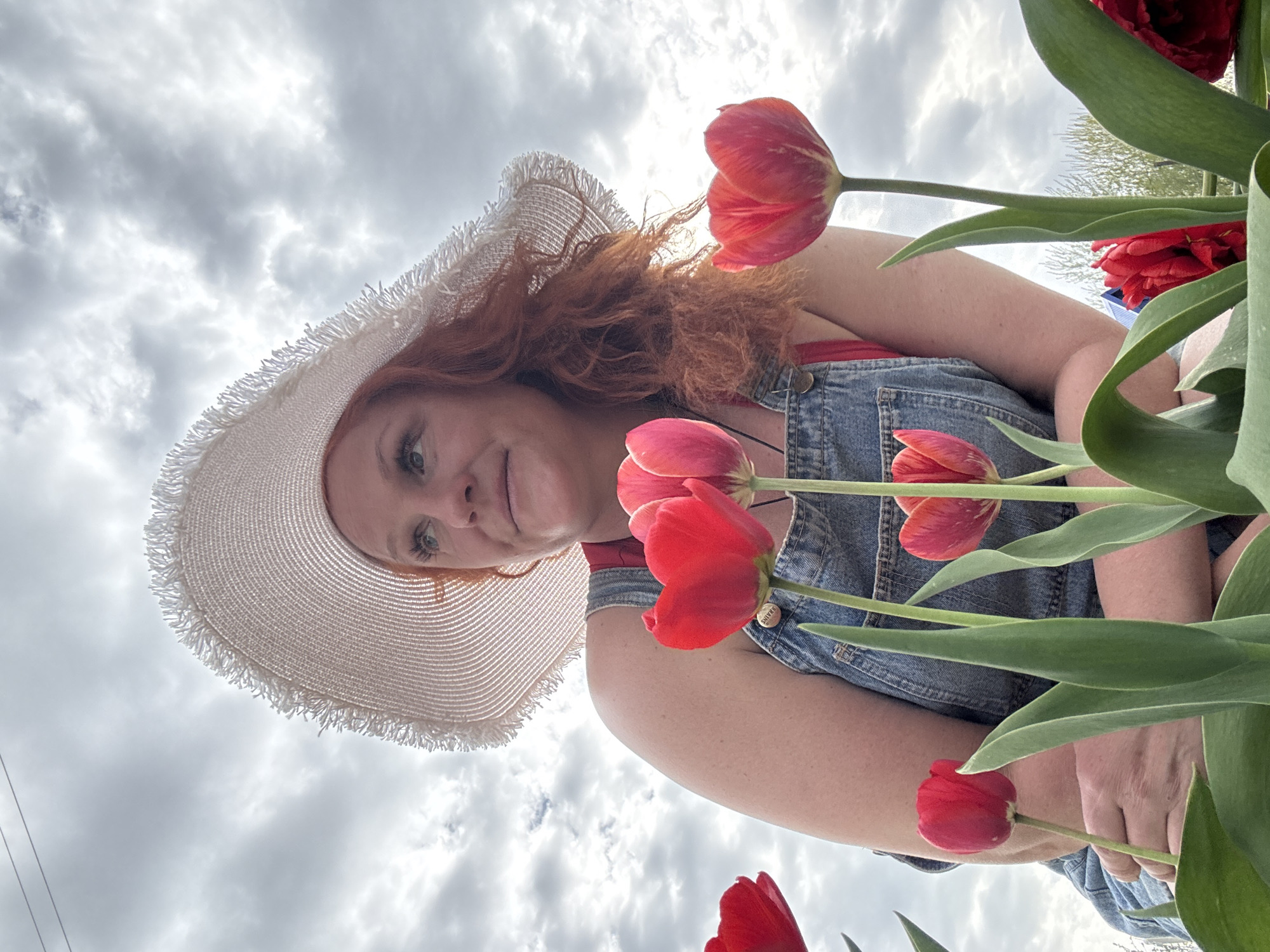 A woman with red hair and a wide-brimmed straw hat is pictured from a low angle, with tulips in the foreground and a cloudy sky as the background. She is wearing denim overalls over a red top. The woman has light skin and blue eyes. The tulips are red, and the scene includes green foliage. The picture was taken in the city of Chaikovsky, Russia.