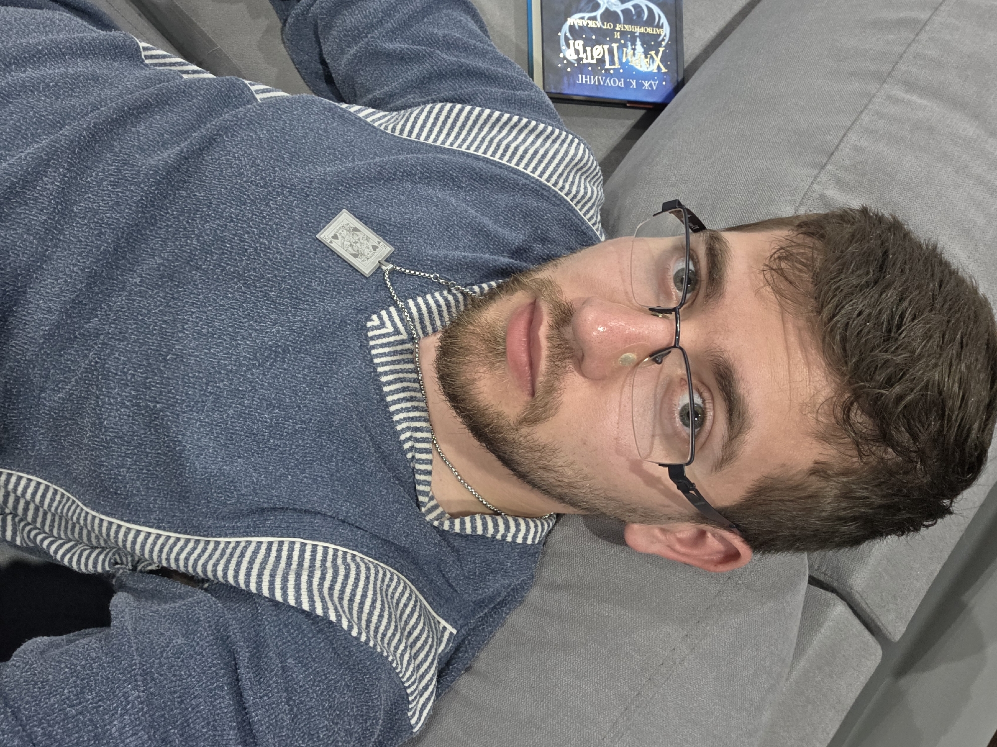 A man with short dark hair and a beard lies on a gray couch, wearing glasses and a blue and white striped sweater with a card pendant around his neck. A book, potentially "Harry Potter and the Prisoner of Azkaban" in Bulgarian, rests nearby. The location is unknown, with no visual cues suggesting Yagodovo, Bulgaria.