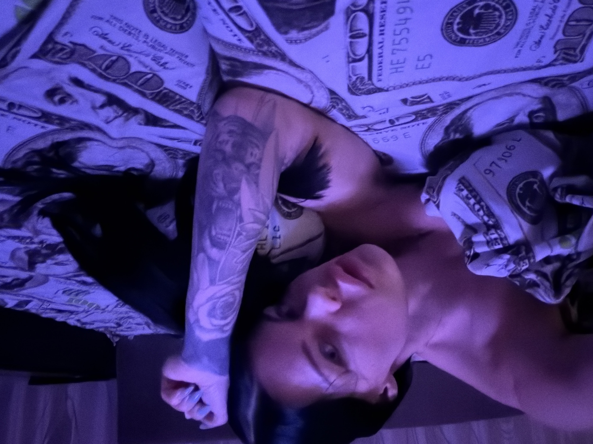 The image shows a woman lying on a bed. The bedsheets are patterned with US one hundred dollar bills. The woman has dark hair and tattoos on her arm. The lighting in the room is predominantly purple. The image does not contain any discernible elements that would allow for its location to be identified within the city of Ufa, Russia.