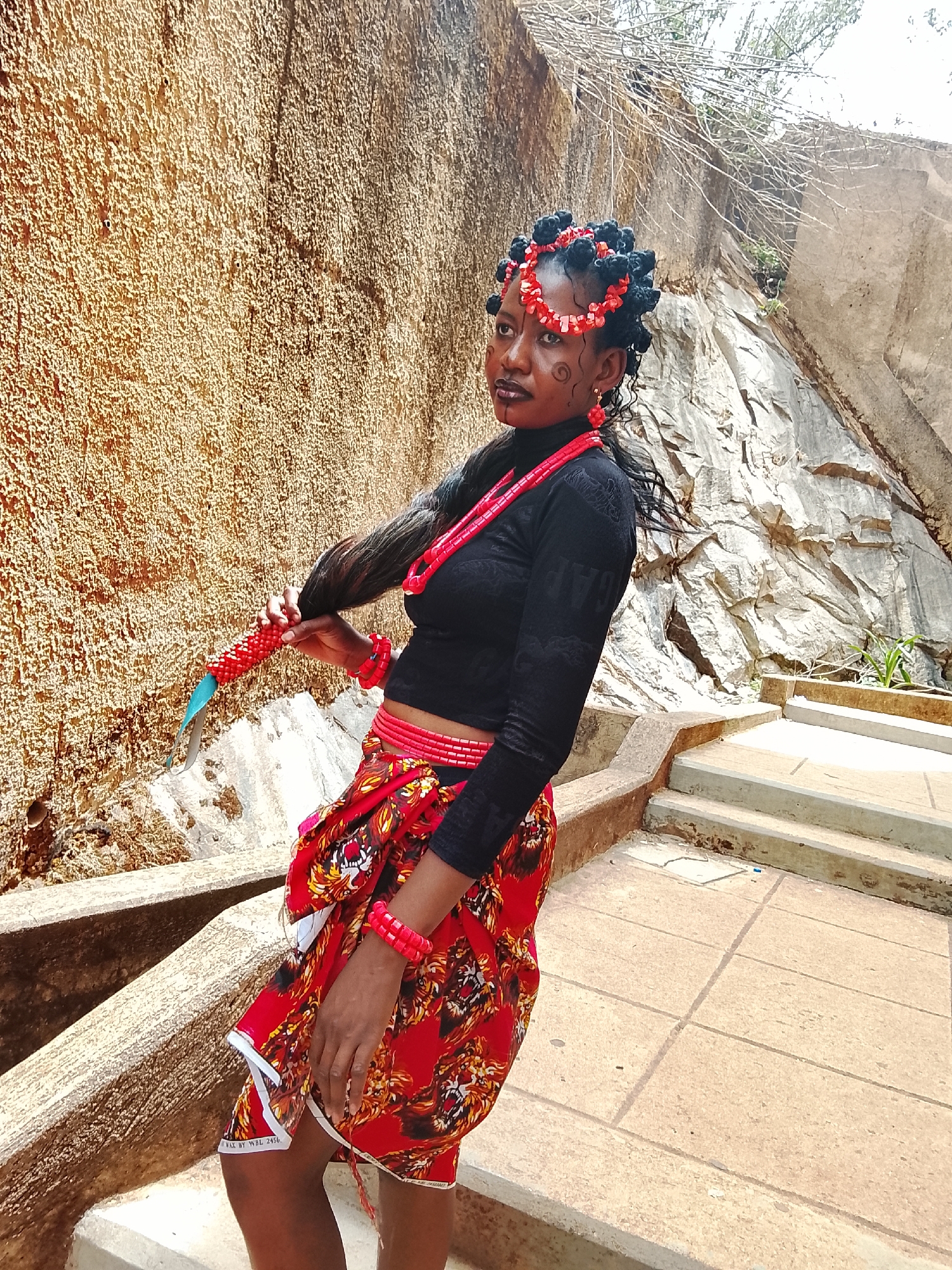 A woman stands on stone steps, likely outdoors, in Jos, Nigeria. She wears a black long-sleeved top, a red patterned skirt with a white hem, and a red beaded necklace and bracelets. Her hair is styled with black knots and red beads. Face paint adorns her cheeks. She holds her long, dark hair, which is wrapped in red beads and a blue fabric, in her left hand. The background includes a textured wall to her left and rocky terrain with stairs to her right. The steps are made of stone tiles.