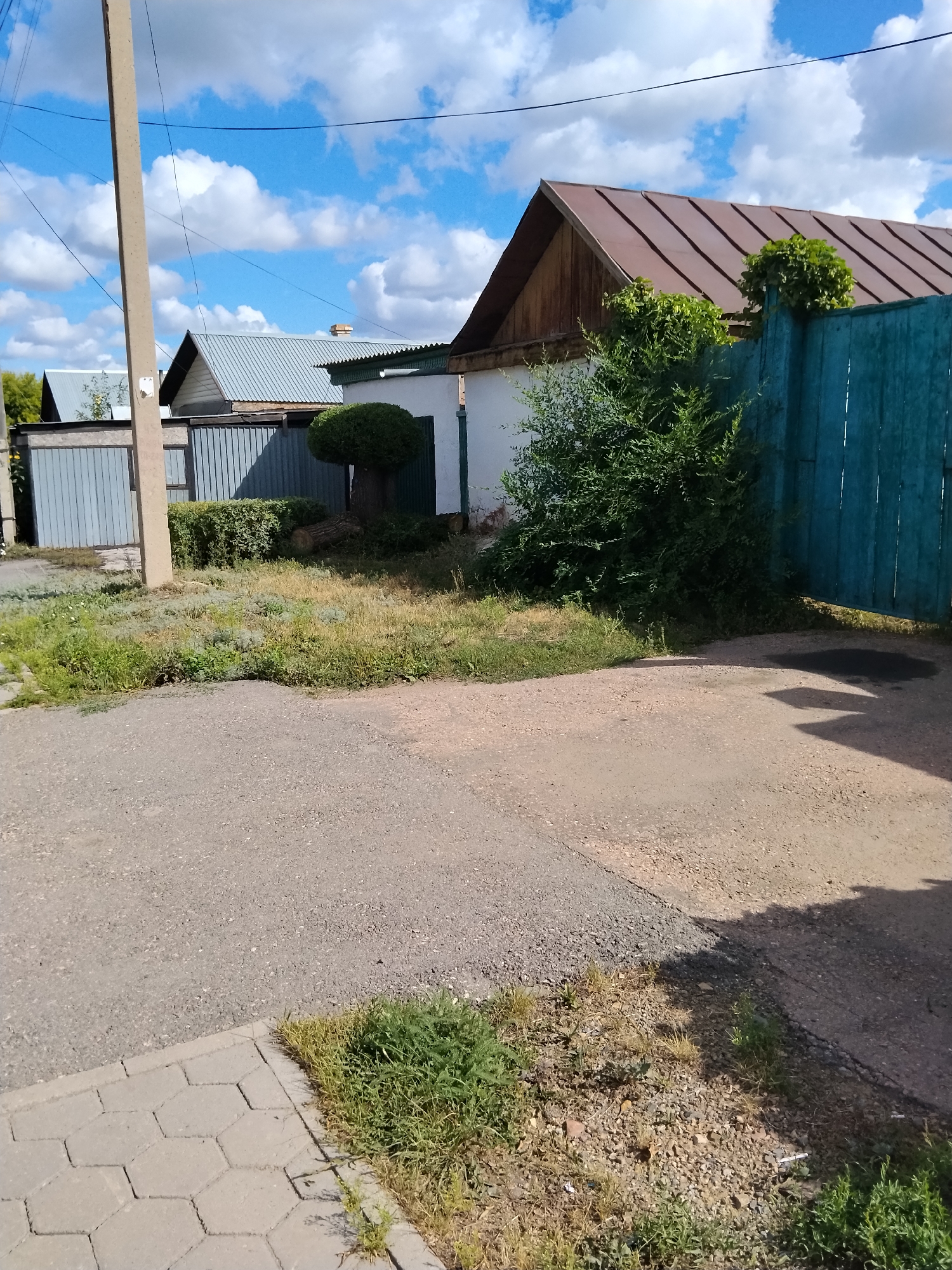 The image shows a residential area in Kokshetau, Kazakhstan. In the foreground, there is a paved area with a sidewalk made of hexagonal pavers on the left. Beyond the sidewalk is a patch of dry grass and gravel. Behind this is a gravel driveway. A concrete utility pole stands on the left side of the frame.
In the middle ground, a light-colored building with a brown metal roof is visible, with a dark green, partially overgrown fence to its right. A large, bushy green shrub is in front of the fence, and a trimmed tree with a rounded top is to the left of the shrub. A felled tree trunk lies on the ground near the trimmed tree. To the left of this building, there is a gray corrugated metal fence and a similar building with a silver metal roof.
The background features a bright blue sky with scattered white clouds. The overall scene depicts a sunny day in a suburban or rural setting. There are no people or animals visible in the image.