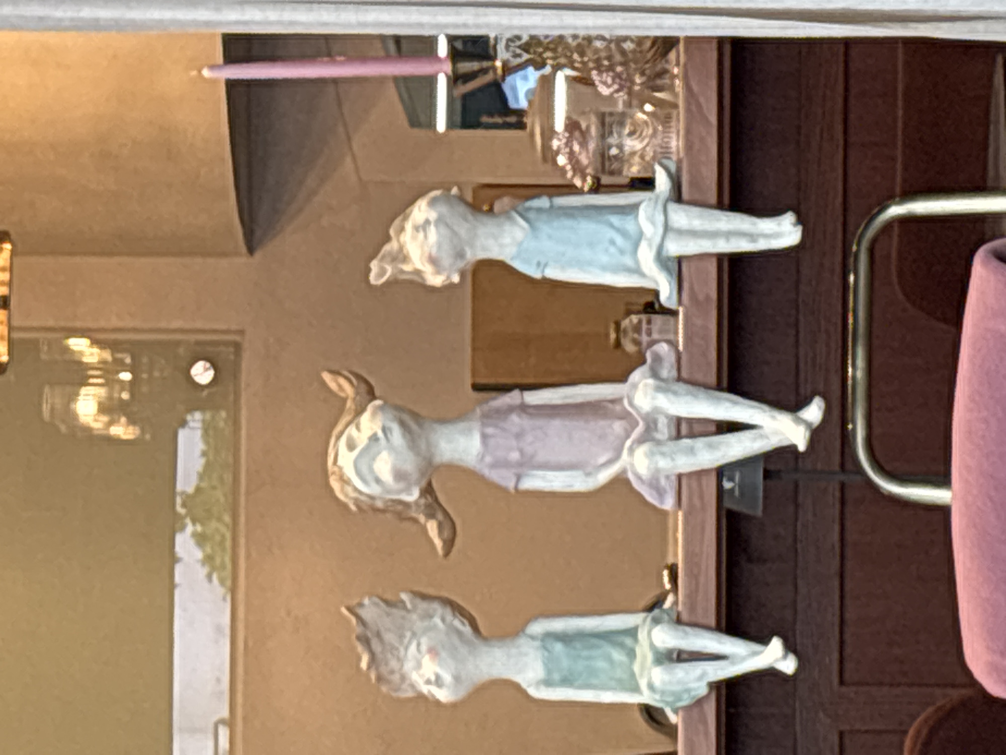 Three ceramic figurines of young girls are displayed on a wooden shelf against a blurred background. The figurines are light-colored with subtle pink blush on their cheeks. From left to right, the first figurine wears a light teal dress, the second wears a lavender dress, and the third wears a light blue dress. All three are depicted sitting with their legs dangling and looking upwards. Behind the figurines, decorative items including jars and a pink candle in a holder are visible. The scene appears to be a shop window display. The location is identified as Varna, Bulgaria, though a specific establishment is not mentioned.