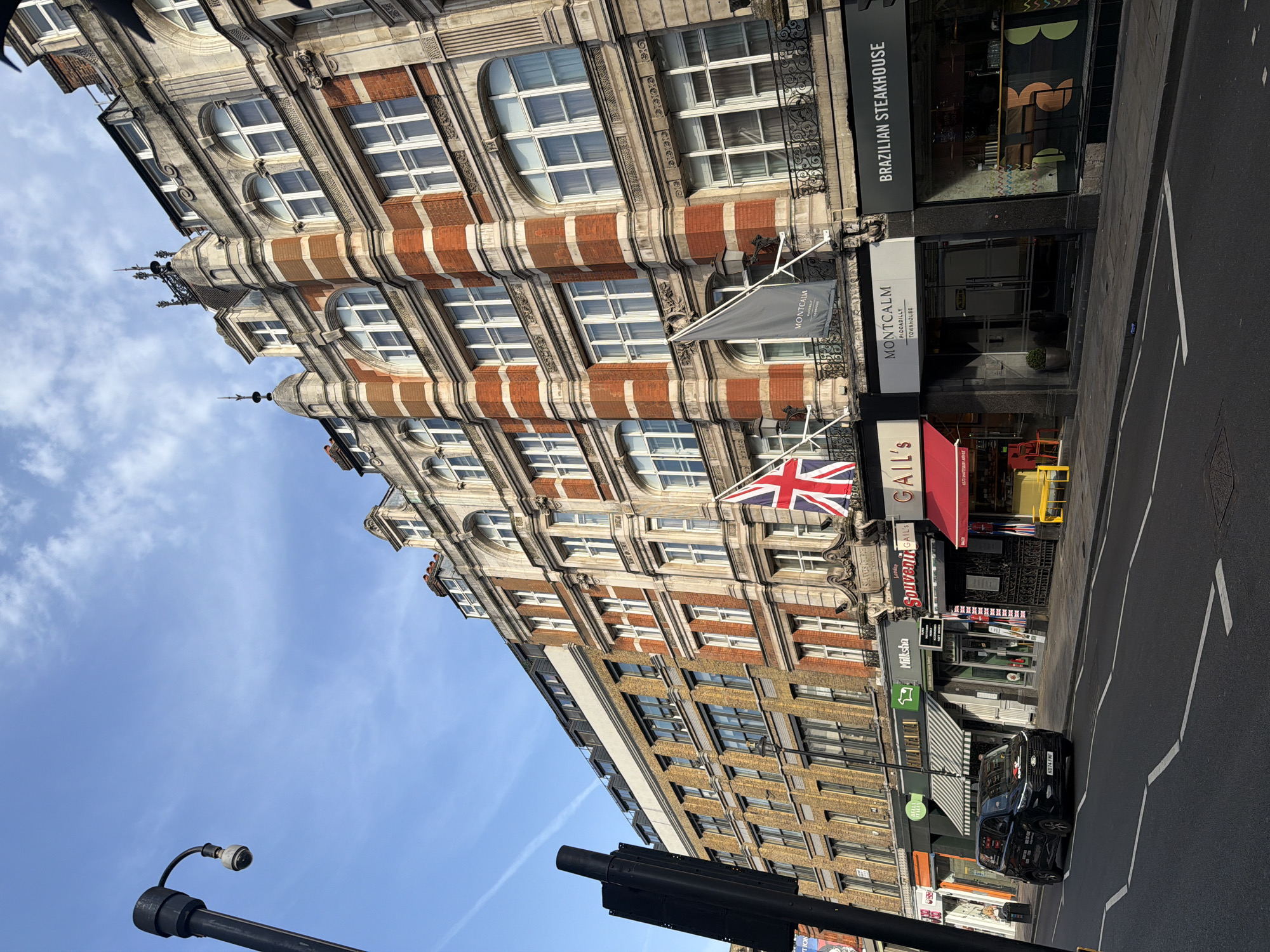 The image shows a street scene in London, United Kingdom, featuring architecture, businesses, and vehicles. The focal point is a tall, ornate building with a combination of red brick and light stone or concrete facades, with distinctive arched windows on upper floors. The building displays flags, including a Union Jack and a flag with "Montcalm" printed on it, suggesting it may be associated with the Montcalm hotel group.
Several businesses are visible at street level, including a "Brazilian Steakhouse," "Gail's," and another identified as "Milksha." Storefronts are adorned with awnings and signage, and there are visible indications of retail activity.
A black car with the license plate "K074 FMF" is parked on the side of the road. In the lower part of the frame, a street lamp or traffic signal pole is positioned, and blue skies are visible above the buildings. Text visible in the OCR data also reveals that it's Piccadilly, Shaftesbury Avenue.