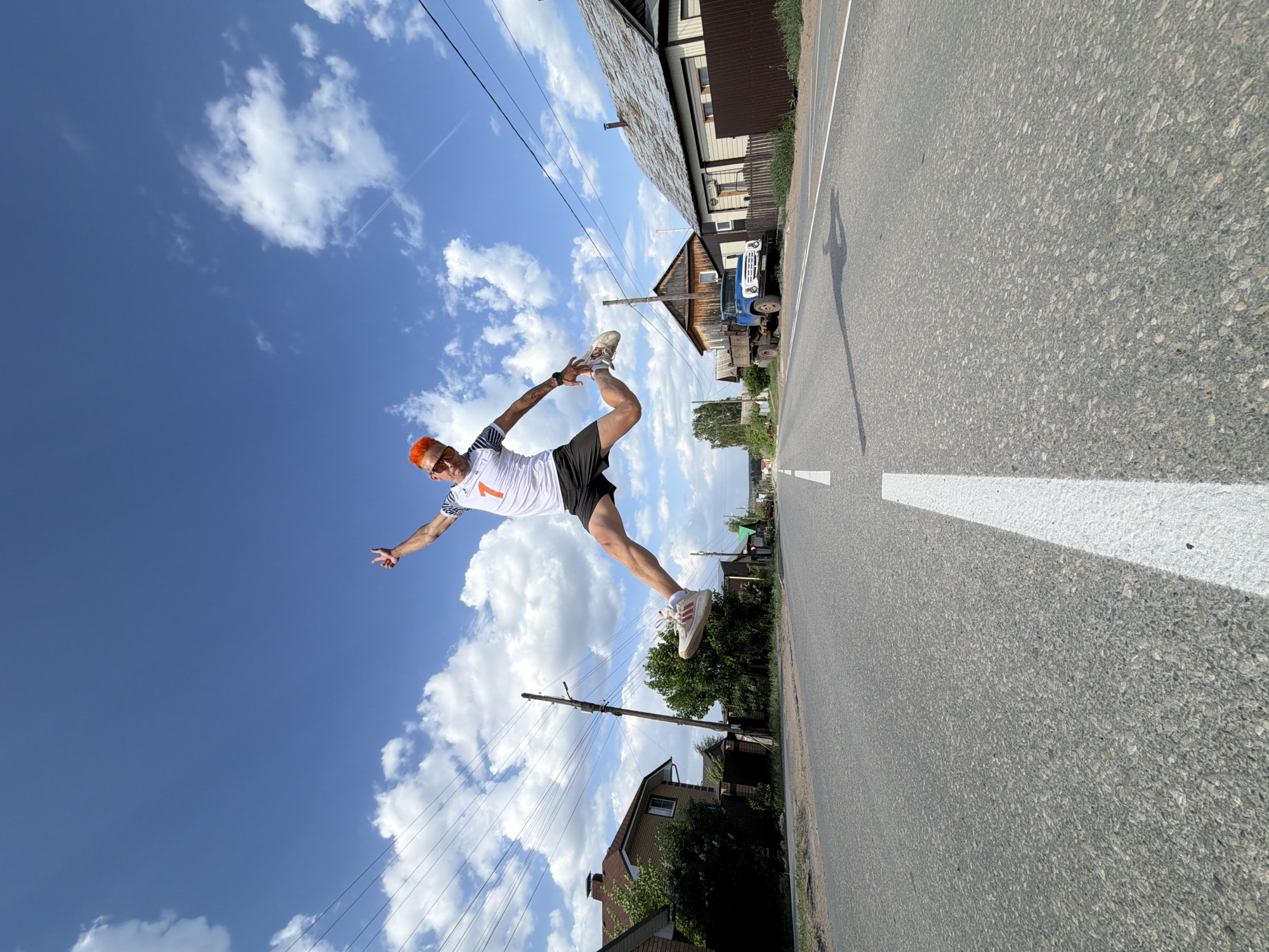 A man with orange hair and sunglasses wearing a white shirt with a large number 1 printed on the front, black shorts, and white sneakers is captured mid-air in a dynamic pose. He holds one foot up near his head while extending the opposite arm, with a hand gesture towards the sky. The scene is outdoors on an asphalt road with a white center line marking. The background features a clear blue sky with scattered white clouds. Power lines stretch across the sky. A truck is visible beside the house on the upper right of the shot.