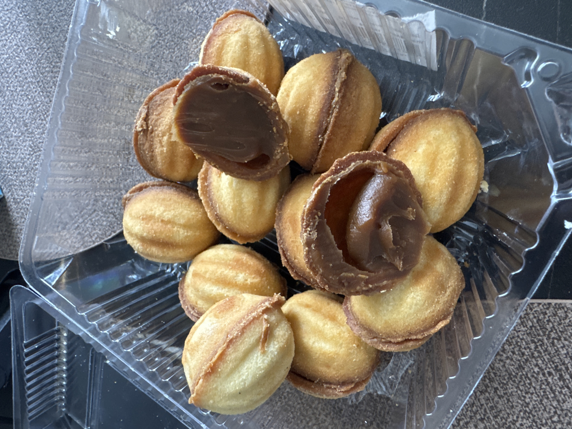 A top-down view depicts a clear plastic container holding approximately a dozen nut-shaped pastries. The pastries are golden brown with ridged surfaces and are filled with a dark brown filling. Two of the pastries are broken open, revealing the filling. The container rests on a surface with a patterned, possibly carpeted or textured, appearance. The image does not reveal a location in Vladivostok, Russia or any other information about a city. No people are visible.