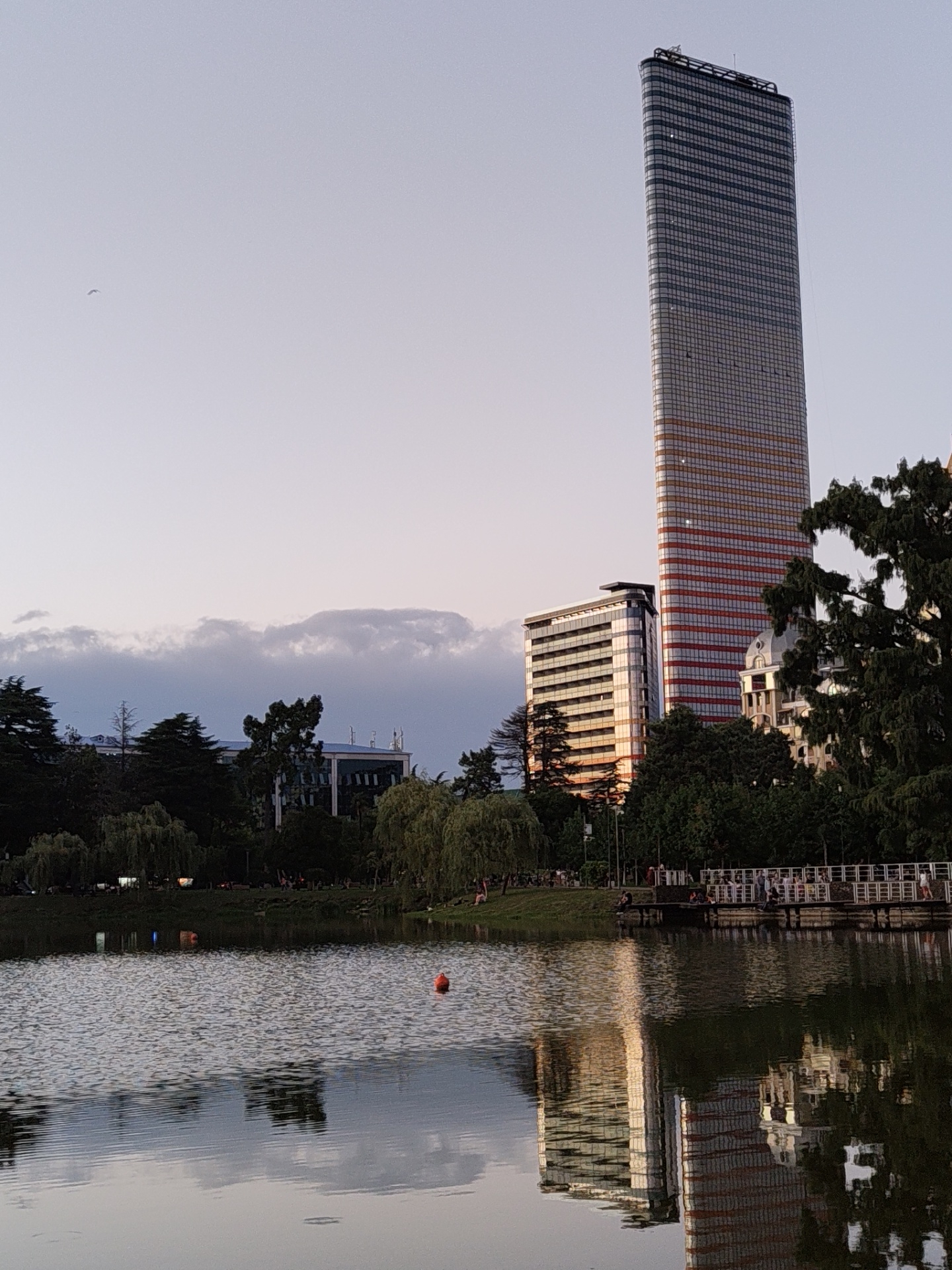 The image depicts a serene lake scene in Batumi, Georgia, with the iconic Batumi Tower and a smaller modern building dominating the skyline. The sun is setting, casting a warm glow on the buildings and reflecting on the calm water. Trees line the lake's edge, with a wooden pier and a railing in the foreground where people are seen strolling and sitting. Two orange buoys float on the lake, adding a touch of color to the tranquil water. The sky is a soft gradient of blue and pink, with a few wispy clouds adding texture. A bird can be seen soaring in the distance, adding to the peaceful atmosphere. The scene captures a moment of urban tranquility, where modern architecture and natural beauty coexist harmoniously.