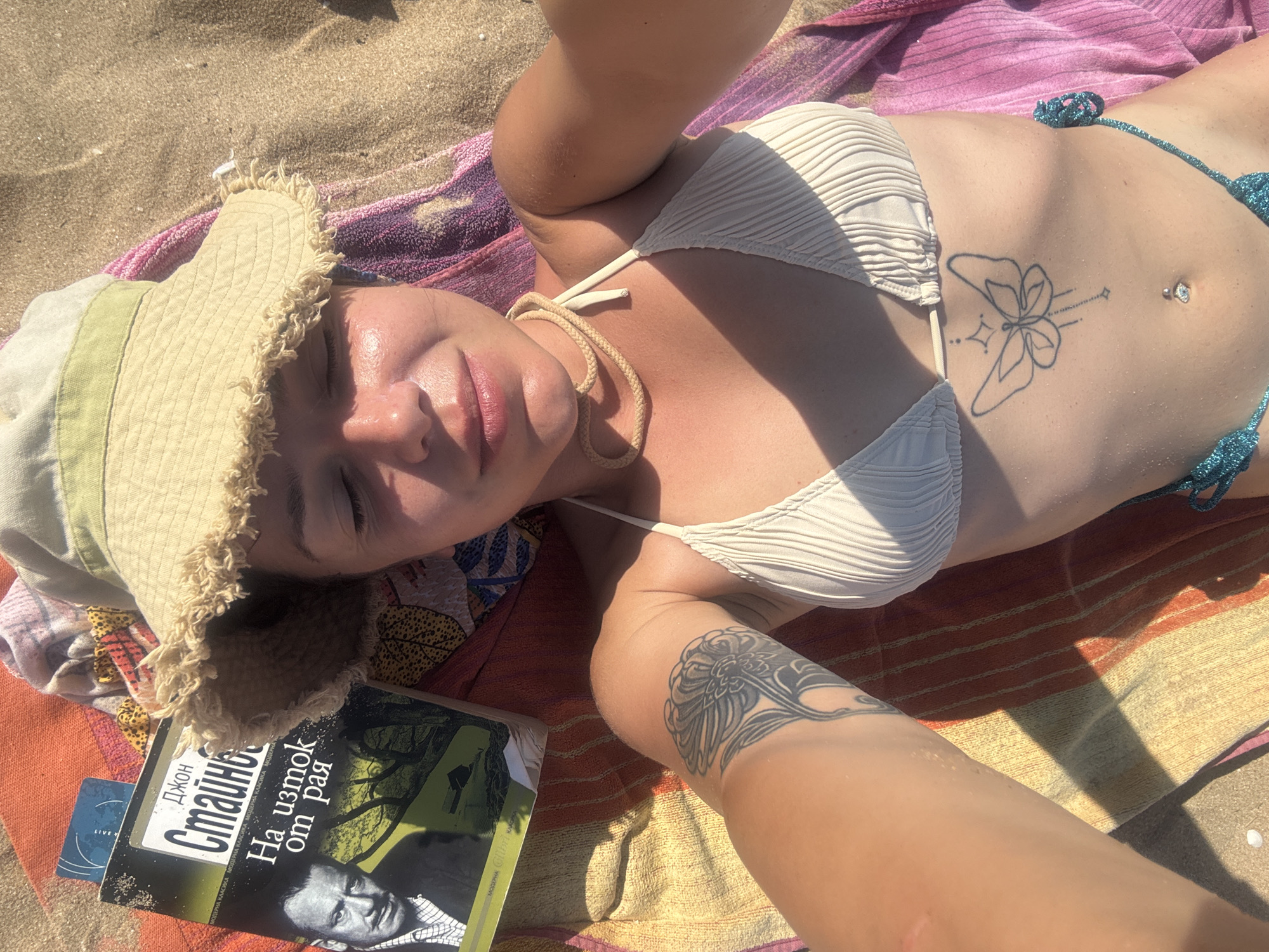 A woman lies on a sandy beach in Sozopol, Bulgaria, basking in the sun on a colorful striped towel. She is wearing a light-colored ribbed bikini top and a floppy hat with a frayed brim. A book titled "На изток от рая" (On the East of Eden) by John Steinbeck is placed near her. She has a butterfly tattoo on her abdomen and another tattoo on her thigh. The sun is bright, casting shadows on the sand and her body, suggesting it is daytime. The overall scene evokes a sense of relaxation and leisure on a warm day at the seaside.