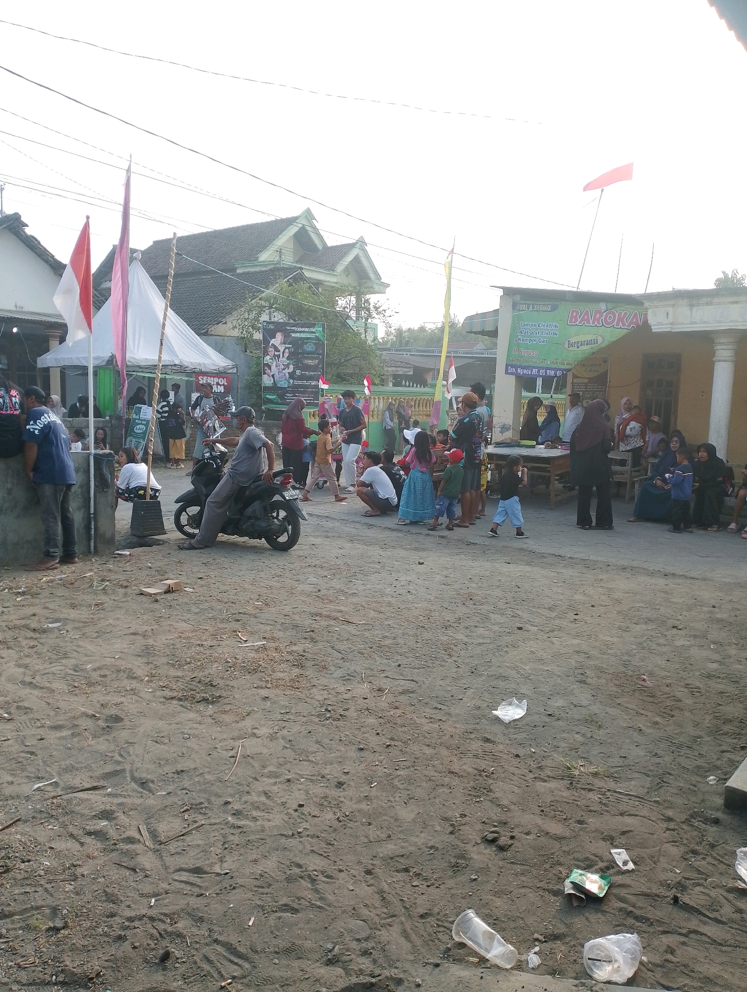 The image depicts an outdoor scene, likely a community gathering or event, in an area with sandy ground, suggesting it could be near a beach or a dirt road in Rembang, Indonesia. In the foreground, the sandy ground is littered with debris, including plastic bottles, wrappers, and twigs. A few people are visible in the mid-ground, some standing and observing, while others are seated. A man in a gray shirt and pants is sitting on a dark-colored motorcycle, which is parked on the sand.
In the background, a tent is set up, adorned with Indonesian flags, indicating a festive or commemorative occasion. Several people, including adults and children, are gathered around the tent and nearby structures. Some individuals appear to be interacting, possibly engaging in conversations or activities related to the event. A banner with text, possibly advertisements or event information, is displayed on one of the buildings. The overall atmosphere suggests a casual, community-focused gathering under a bright sky.