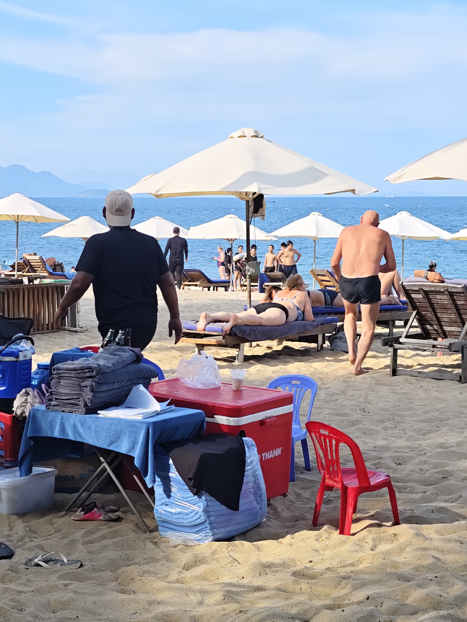 The image depicts a busy beach scene in Nha Trang Ward, Vietnam. In the foreground, a man in a black t-shirt and a baseball cap walks away from the camera. He is near a table covered with a blue cloth, on which several folded towels and some papers are placed. Two bottles of drink are visible on the table. Next to the table are two large red coolers, a blue chair, and a stack of towels wrapped in plastic. A pair of pink and white flip-flops are on the sand near the folding table. Further back, several beach umbrellas provide shade for lounge chairs occupied by beachgoers. A man in black swim trunks walks away from the camera towards the sea. Another man, also in black swim trunks, is seen walking towards the sea in the background. A woman in a bikini is lying on a lounge chair. The sea is visible in the background, with a few boats on the horizon. The sky is blue with some scattered clouds. The sand is light brown and appears to be typical beach sand.