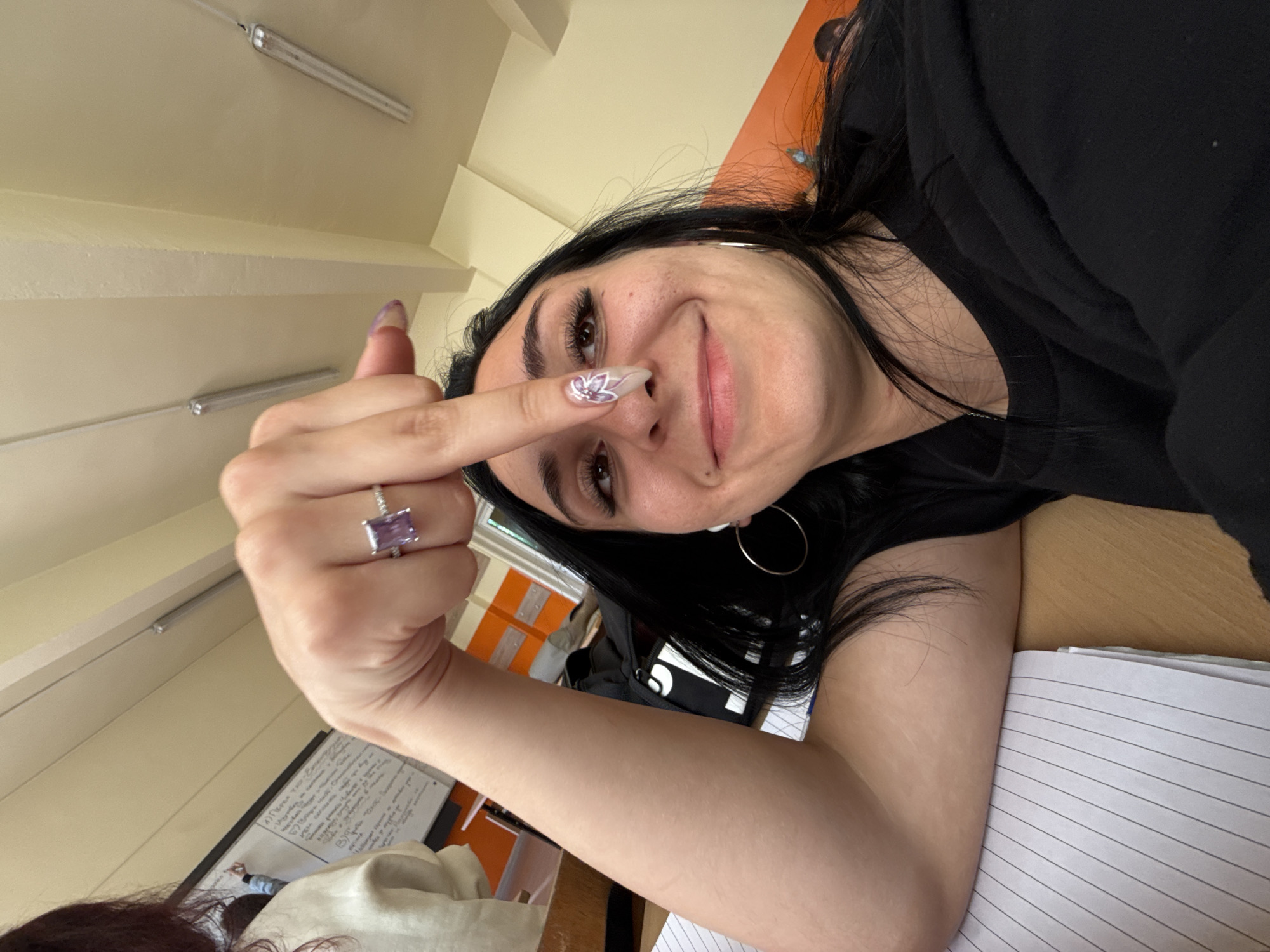 A woman is in what appears to be an indoor classroom, facing the camera at an angle. She is holding up her middle finger. The woman has black hair and wears a black shirt. She has a silver ring on her finger, and a patterned design on her middle fingernail. A notebook and papers sit on a wooden desk. A whiteboard with writing is visible in the background. The location cannot be determined to be in Sofia, Bulgaria from this image.
