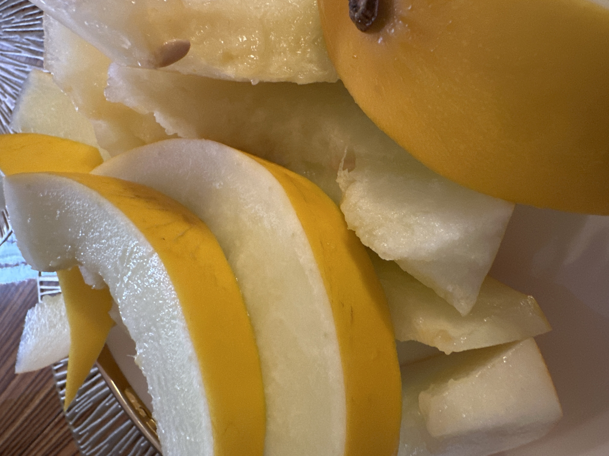 The image shows a close-up view of sliced honeydew melon and possibly some cantaloupe. The melons are cut into wedges and arranged in a bowl. The lighting is bright, highlighting the juicy texture of the fruit. The background is blurred, but appears to be a wooden surface. There are no people or animals visible in the image. The location is Yuzhnouralsk, Russia.