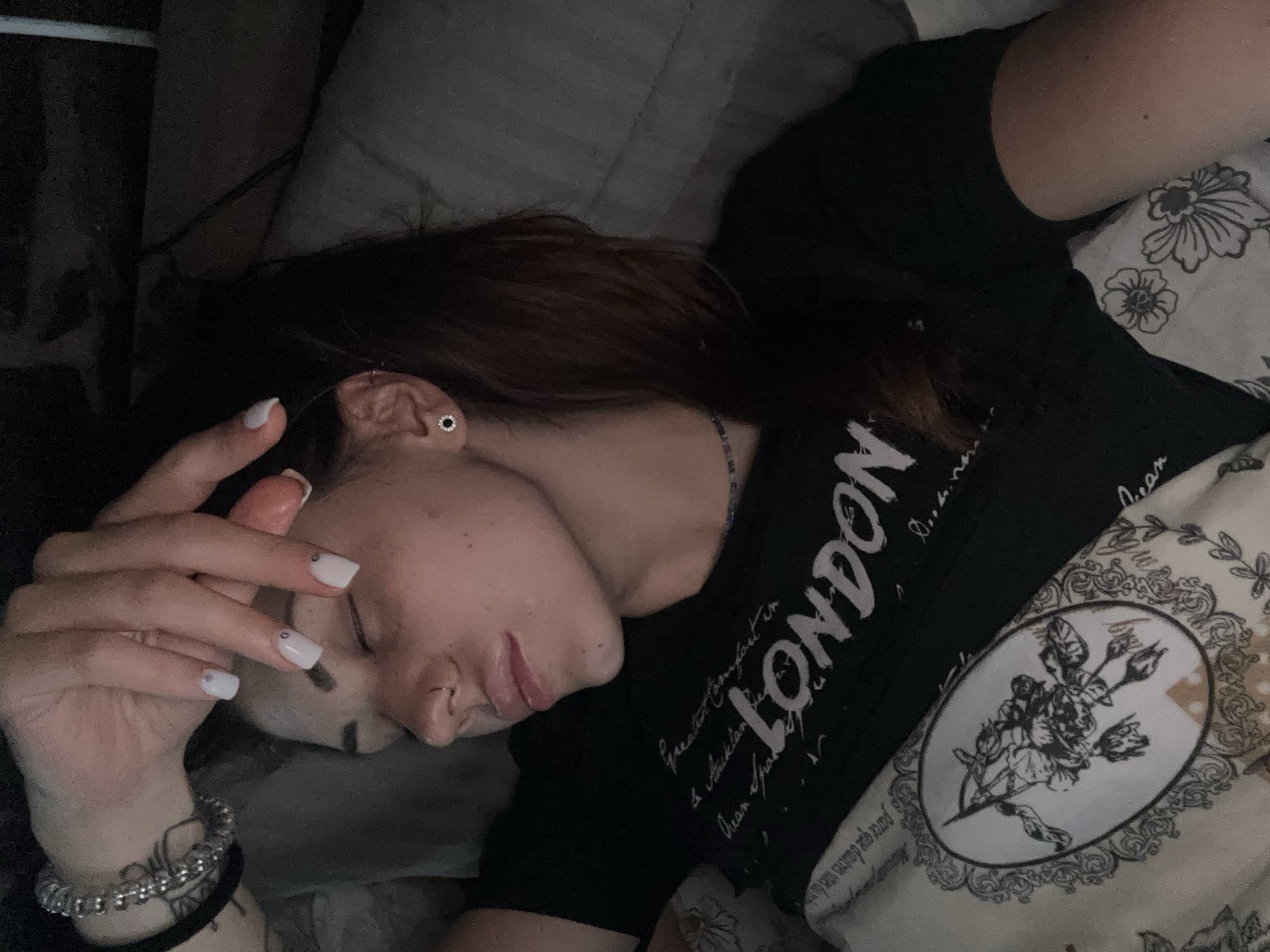 A young woman lies sleeping in a bed. She is turned on her side, facing the camera, with her hand resting near her forehead. She has long brown hair and wears a black t-shirt with "LONDON" printed on it. She has a nose piercing and a stud earring. Her fingernails are painted white with small gem accents. A bracelet is visible on her wrist. The bed has a patterned pillowcase and a gray duvet. The lighting is dim.
There is no location information, Belopoptsi, Bulgaria, in the image.