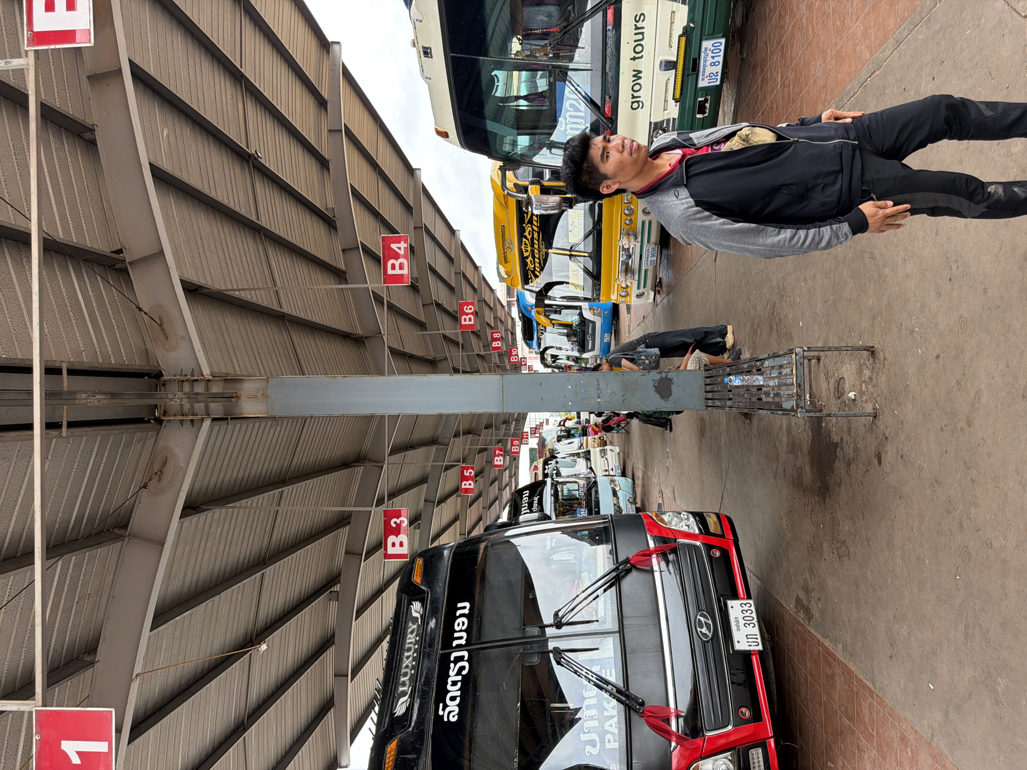 The image depicts a bus station in Phakhao, Laos. In the foreground, a man stands looking to the right. He is wearing a dark grey and black zip-up jacket over a red shirt, and dark pants. Behind him, several buses are parked under a covered shelter. The bus on the left is black with "LUXURY" branding and text in a script. The next bus is blue, followed by a yellow bus with "VIP" and "Mousin" branding. Further back, a white and green bus with "grow tours" written on it is visible. Red signs indicating bus bay numbers (B3, B4, B5, B6, B7, B9, B10, B11, B12, B13) are suspended from the shelter. The ground is paved with concrete and some reddish tiles. The environment appears to be outdoors, with a cloudy sky visible above the shelter. Several other individuals are seated or standing in the background near the buses. The overall scene captures a moment at a busy transportation hub.