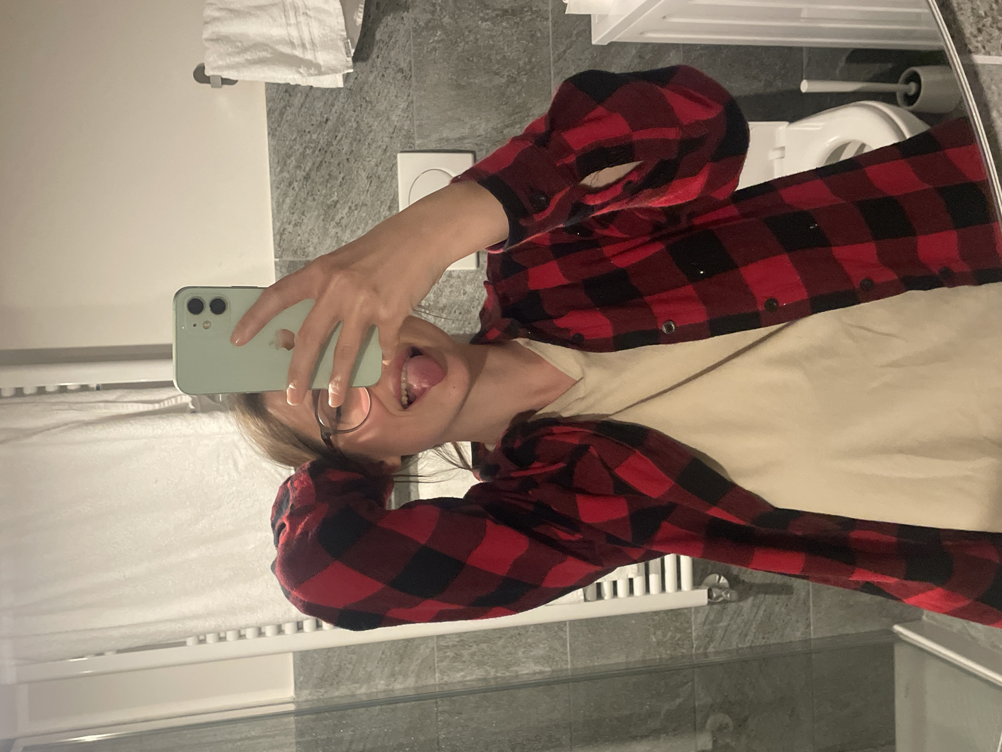 A young woman is taking a selfie in a bathroom. She is wearing glasses and a red and black plaid shirt over a beige top. She is sticking her tongue out at the camera. The bathroom has grey tile, a white towel rack, a toilet, and a white cabinet. The location of the bathroom is not discernible within the city of Sappada / Plodn / Sapade, Italy.