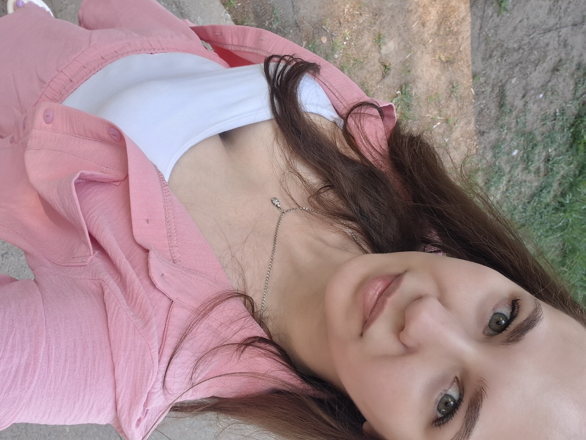 A low-angle, close-up shot features a woman with dark brown hair, fair skin, and striking green eyes. She is wearing a white tank top under a pink button-down shirt. A delicate silver necklace with a small pendant is visible. The background shows a patch of ground with sparse greenery and a glimpse of concrete. The exact location within the city of Chaikovsky, Russia cannot be determined from the visual data provided.